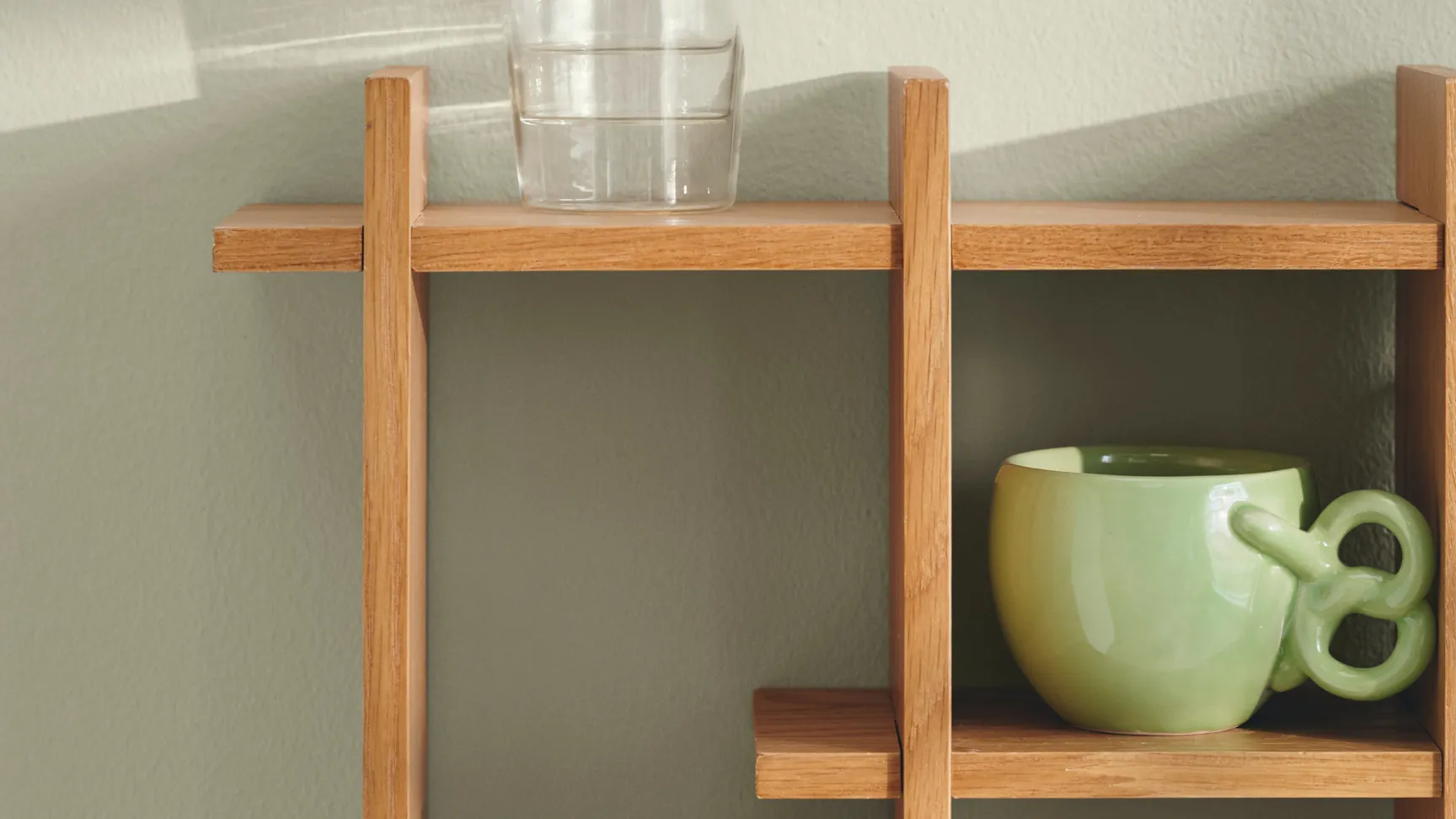Billede af et træhyldesystem med tre klare glas med guldkant øverst, en grøn keramikbeholder til venstre og en metalvase i midten, alle mod en lys grøn væg.