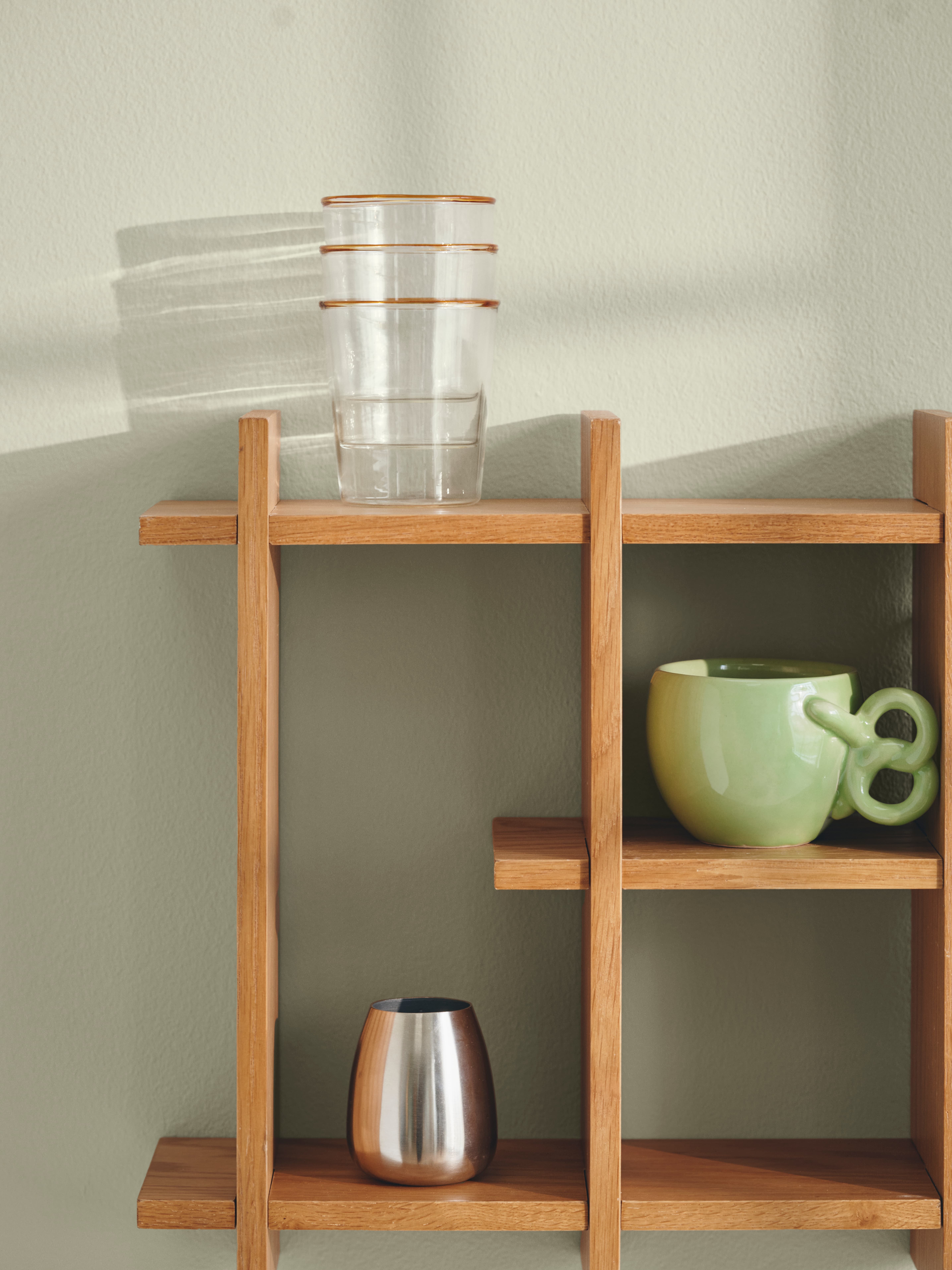 Billede af et træhyldesystem med tre klare glas med guldkant øverst, en grøn keramikbeholder til venstre og en metalvase i midten, alle mod en lys grøn væg.