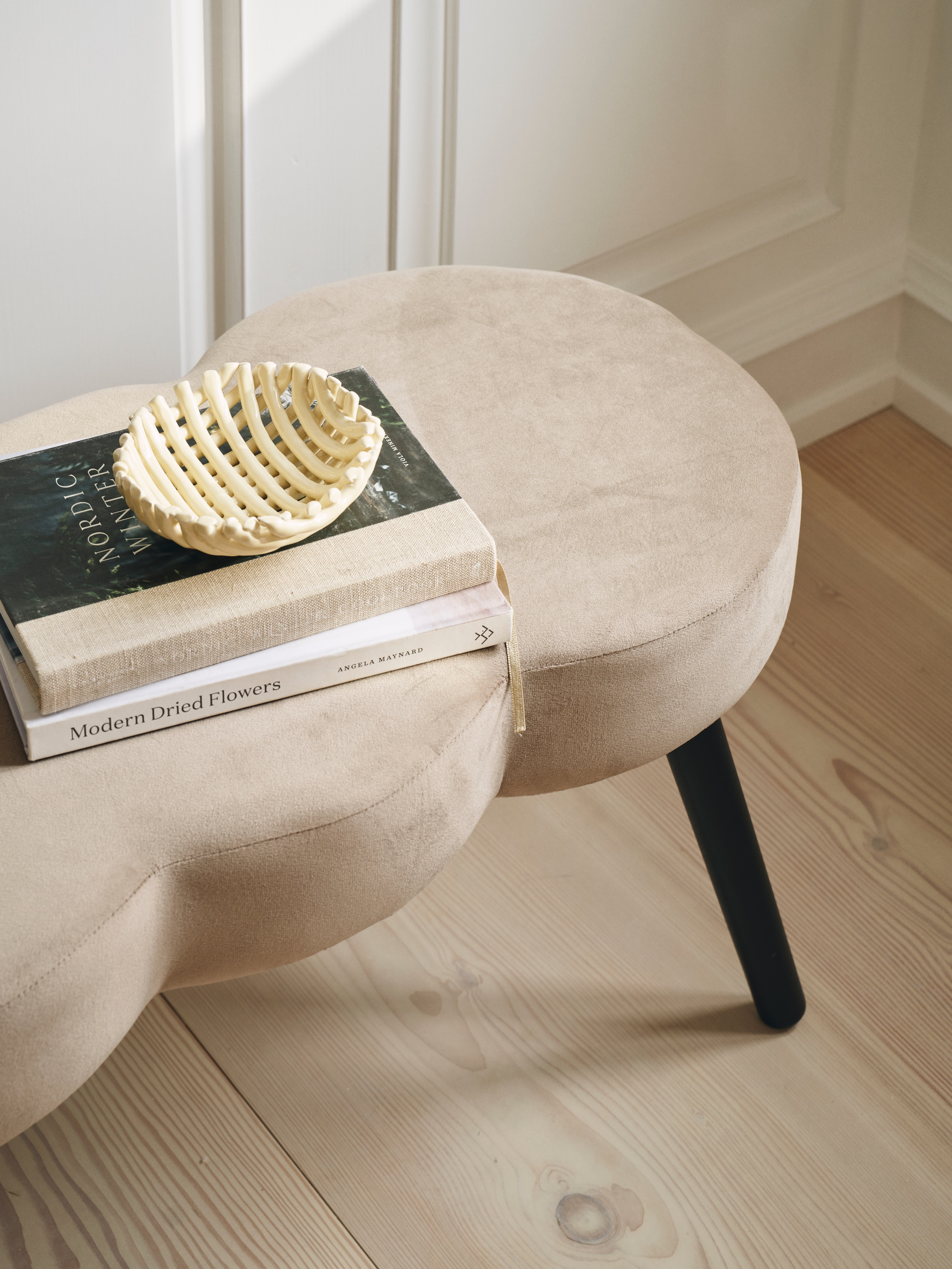 Et lyst, moderne sofabord i beige stof med runde former, dækket af en bog med titlen "Nordic Winter" og en dekorativ skål i lysfarvet materiale. Rummets lyse trægulv og væg skaber en elegant atmosfære.