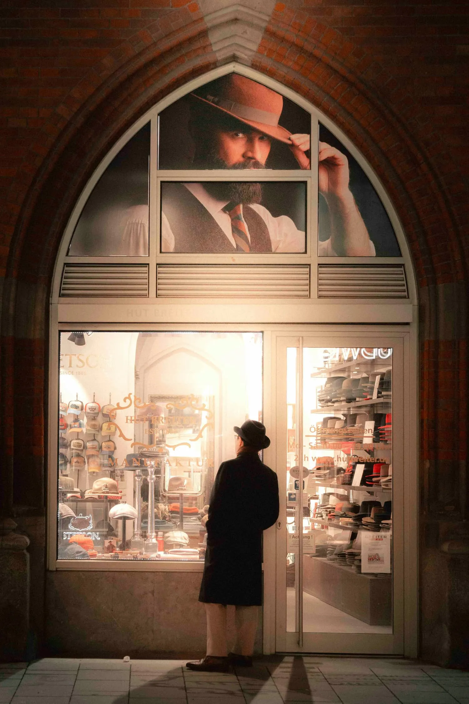 En person i en mørk frakke og hat står foran en butik, der sælger hatte, med en stor plakat af en velklædt mand over vinduet.