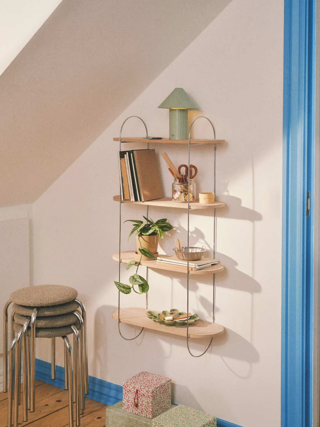 Hyldesystem med tre hylder, et grønt lampe, bøger, planter, og pyntede gaveæsker placeret ved en væg med blå kanter.