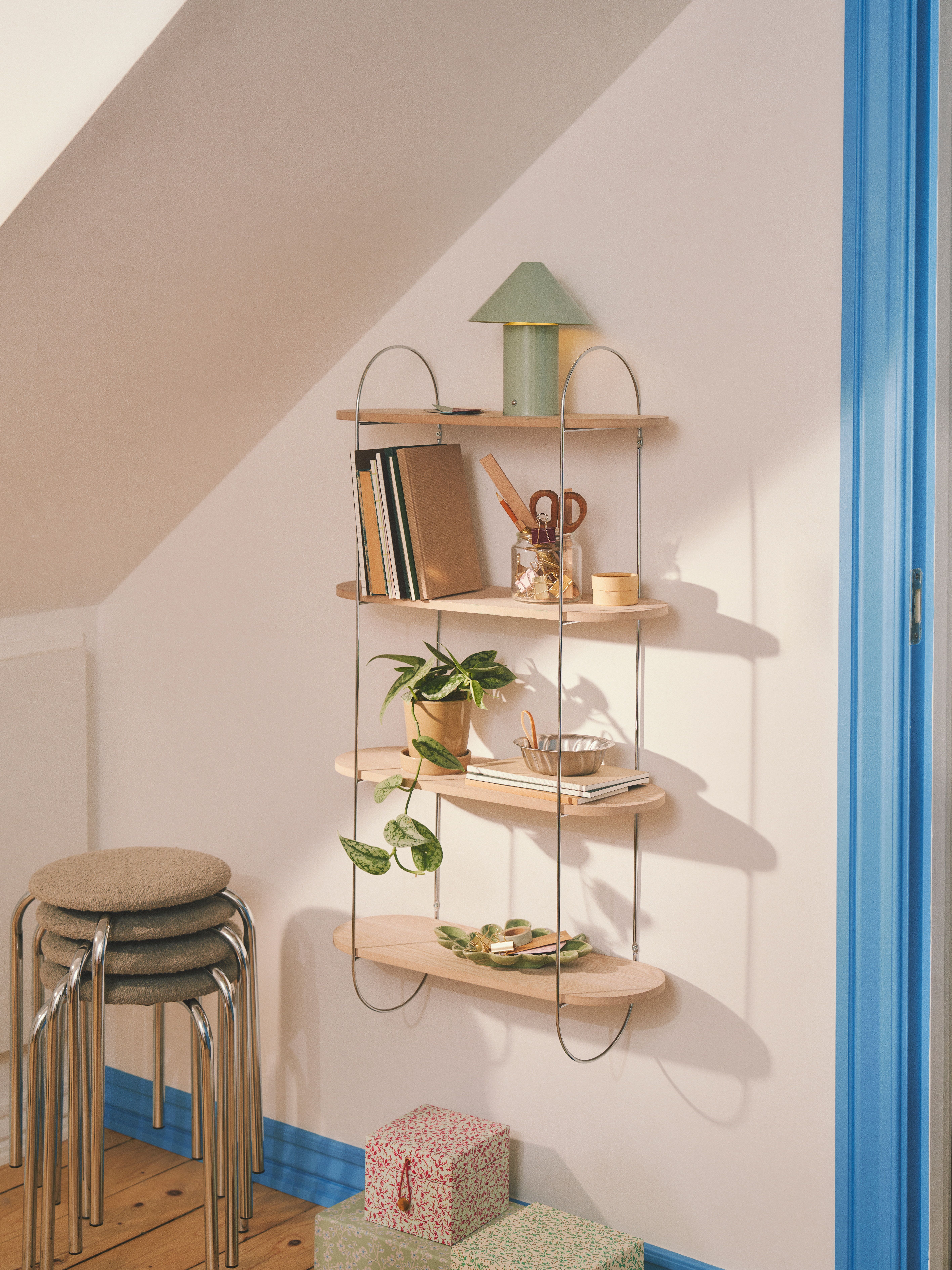 Hyldesystem med tre hylder, et grønt lampe, bøger, planter, og pyntede gaveæsker placeret ved en væg med blå kanter.