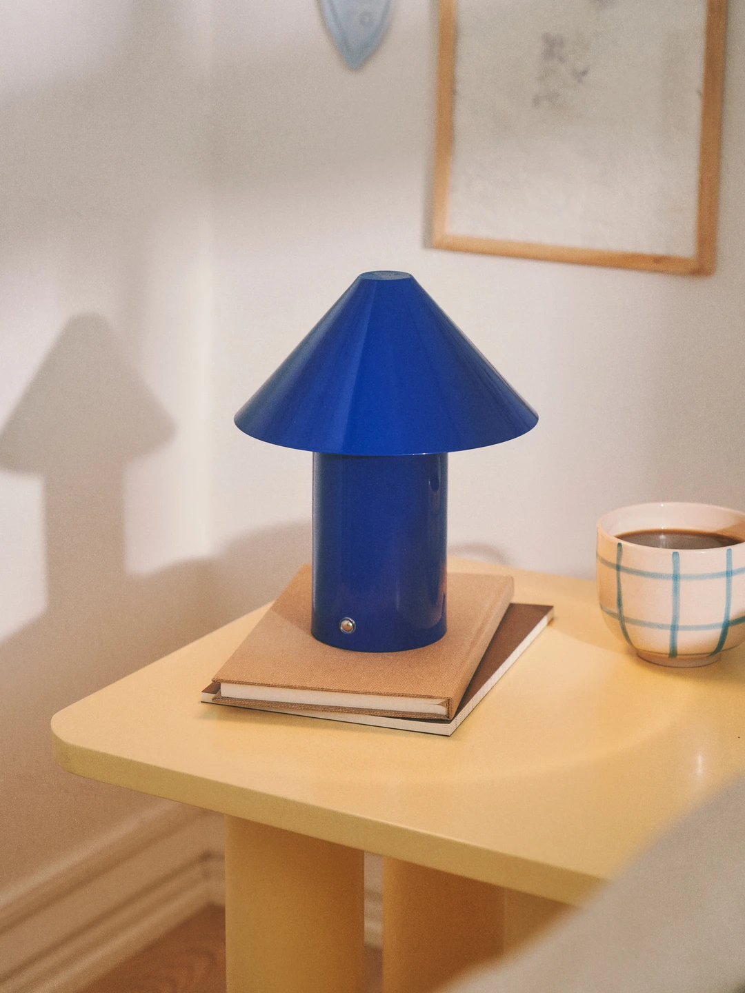 Blå lampe med kegleformet skærm, placeret på en gul bordplade, med bøger under og en kop kaffe ved siden af.