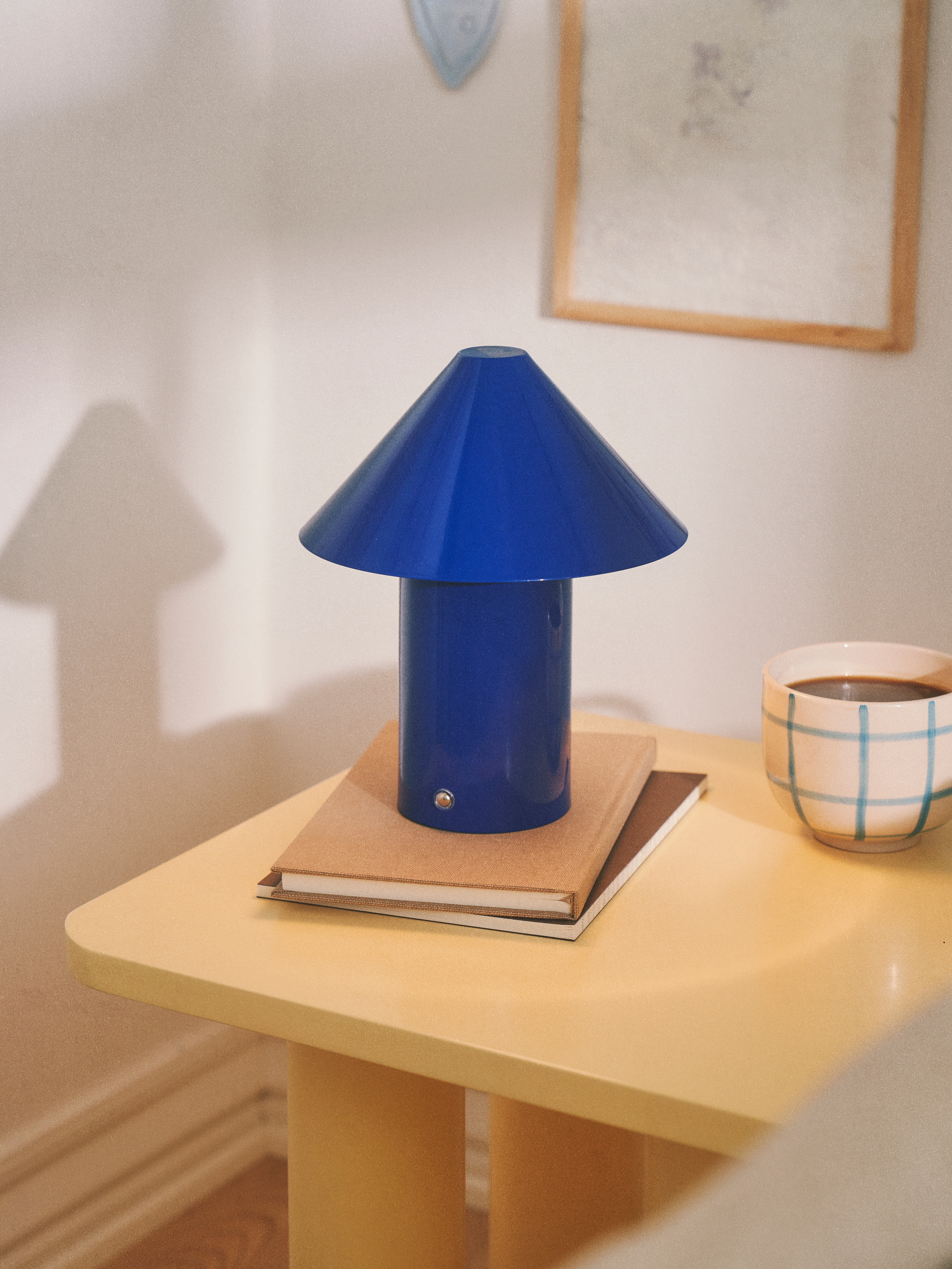 Blå lampe med kegleformet skærm, placeret på en gul bordplade, med bøger under og en kop kaffe ved siden af.