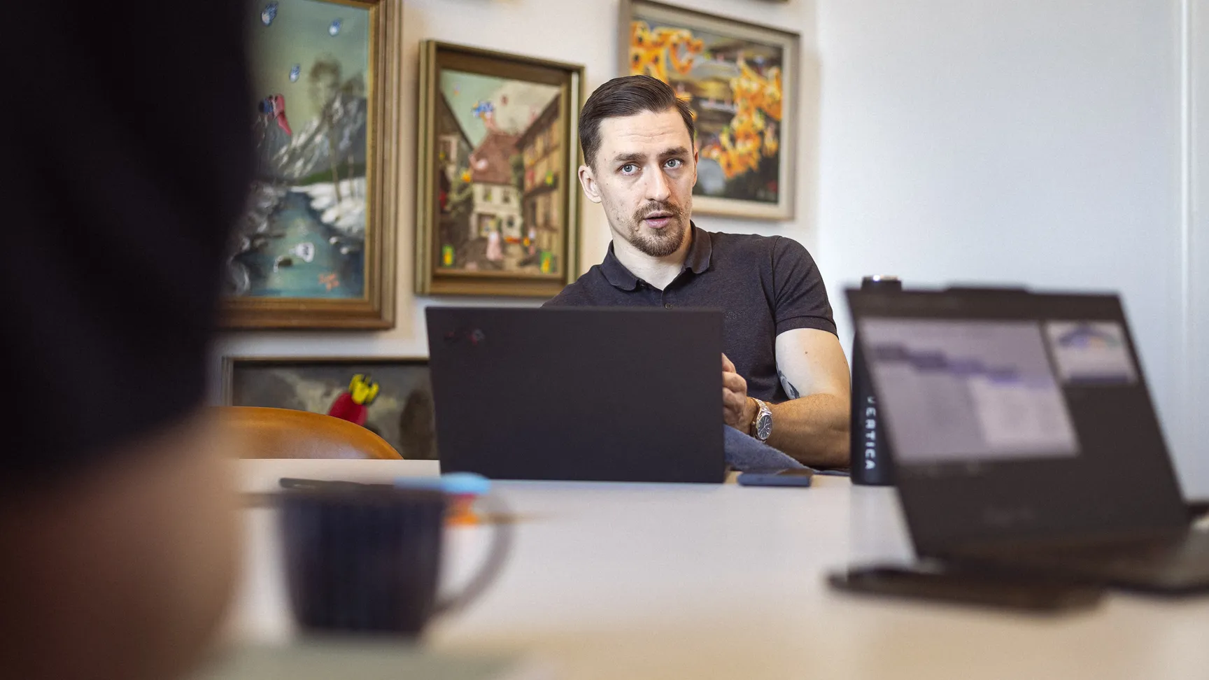 A focused professional in a polo shirt engaged in a discussion during a meeting, with a laptop and a coffee cup on the table, surrounded by colorful artwork on the walls.