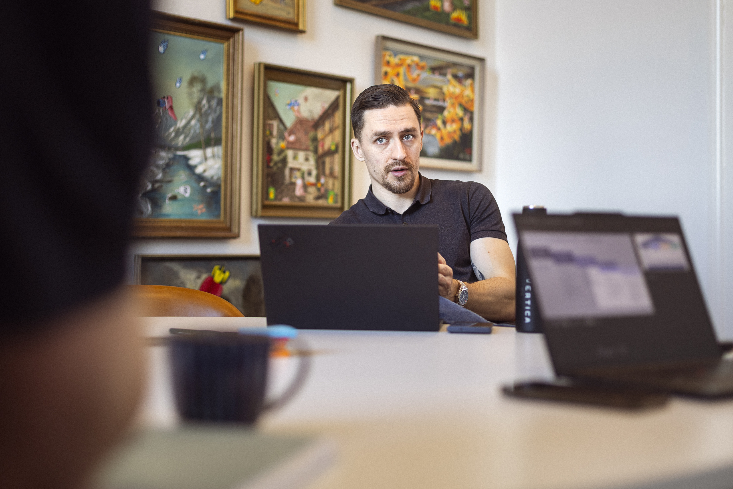 En mand sidder ved et bord med en bærbar computer, mens han taler. I baggrunden er væggen dekoreret med farverige kunstværker, og en kaffekop er synlig på bordet.