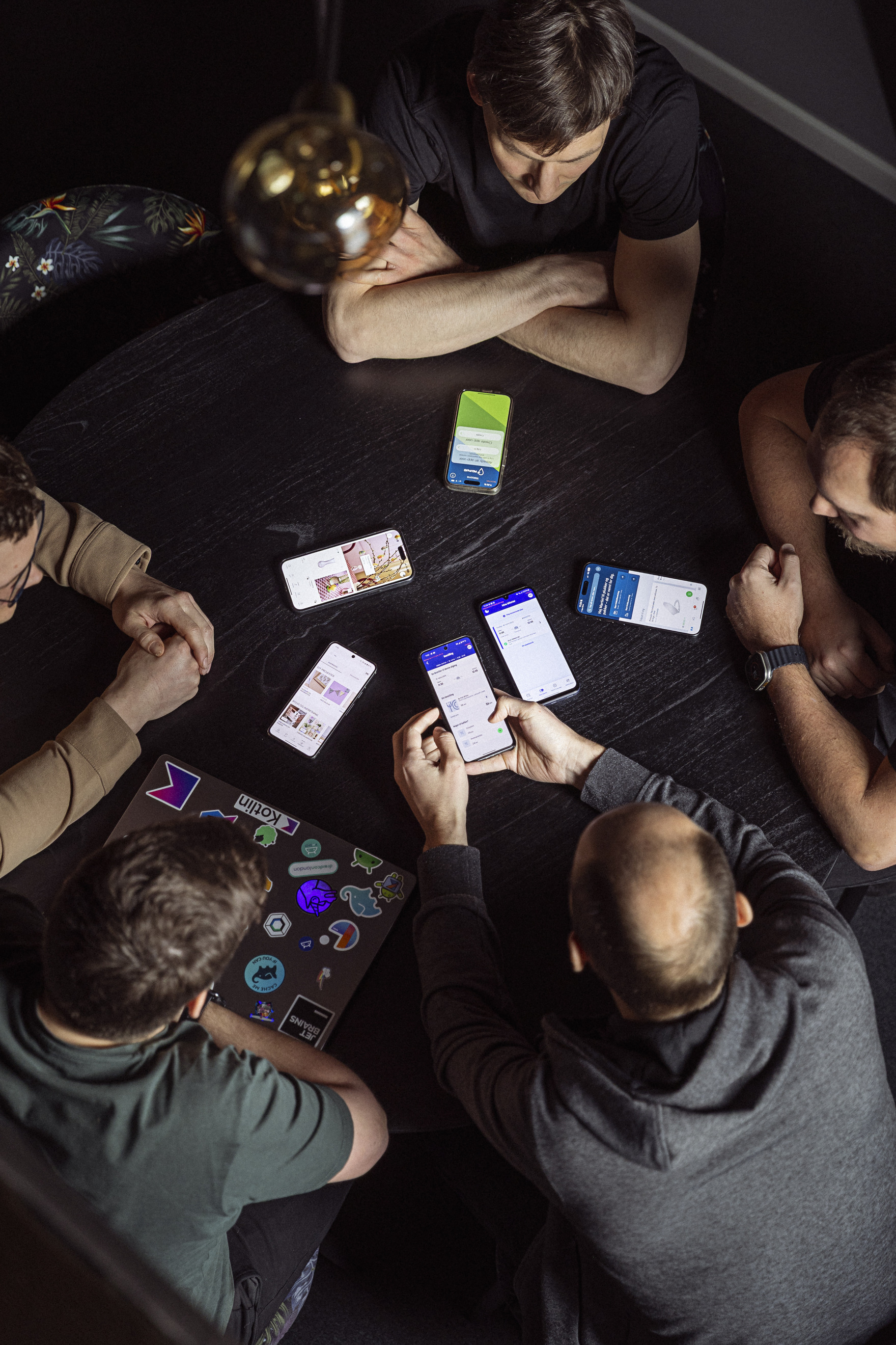 Gruppediskussion ved et bord med fem personer, der ser på deres smartphones og en laptop med klistermærker, i et moderne rum med dæmpet belysning.
