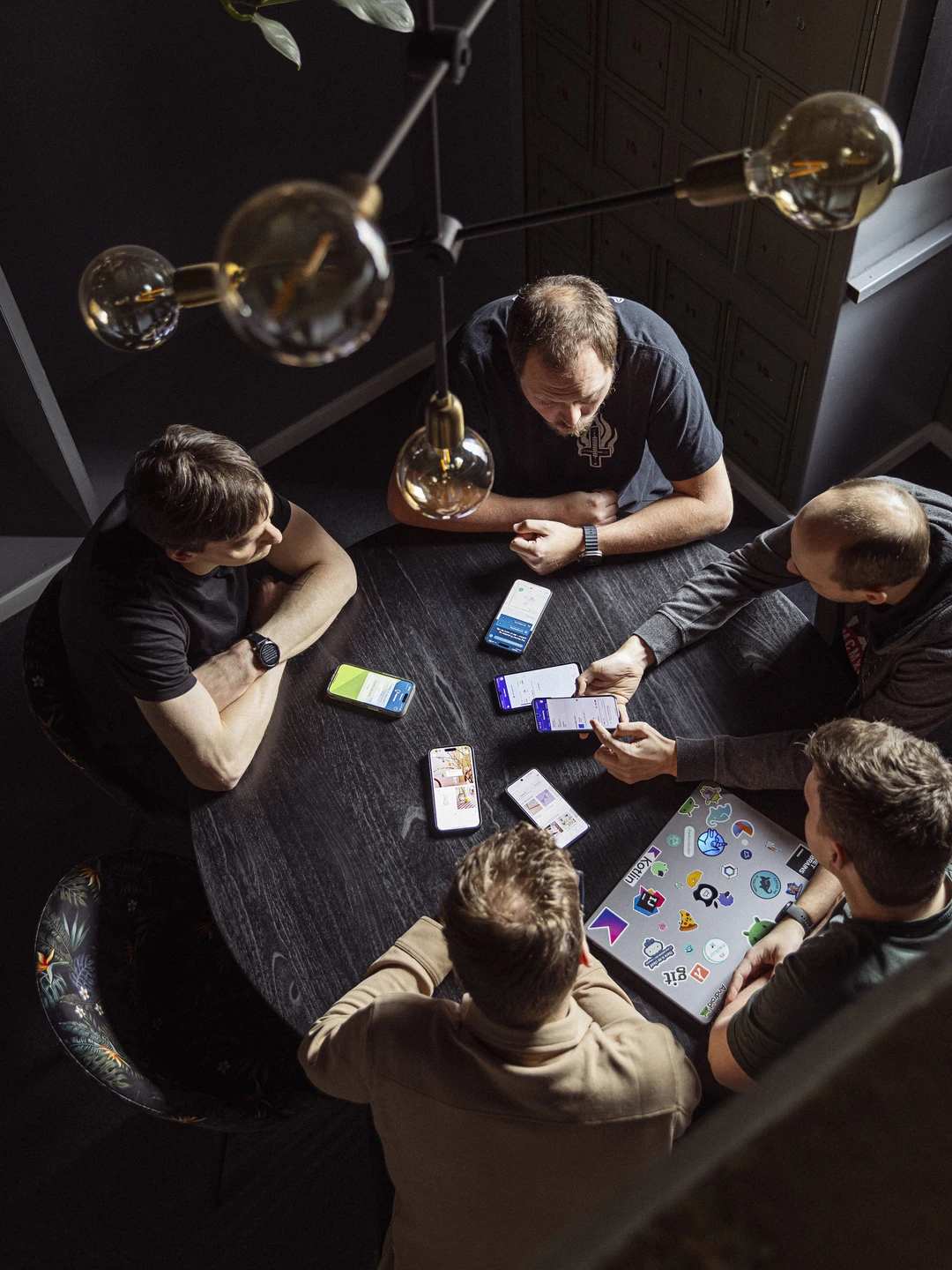 Gruppe af mennesker, der sidder ved et rundt bord, diskuterer og viser indhold på smartphones og en laptop, omgivelsene har en moderne stil med grønne planter og designerbelysning.