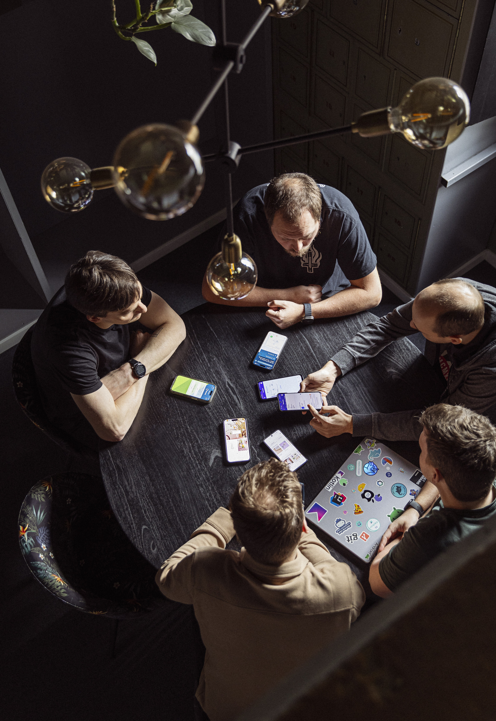 Gruppe af mennesker, der sidder ved et rundt bord, diskuterer og viser indhold på smartphones og en laptop, omgivelsene har en moderne stil med grønne planter og designerbelysning.