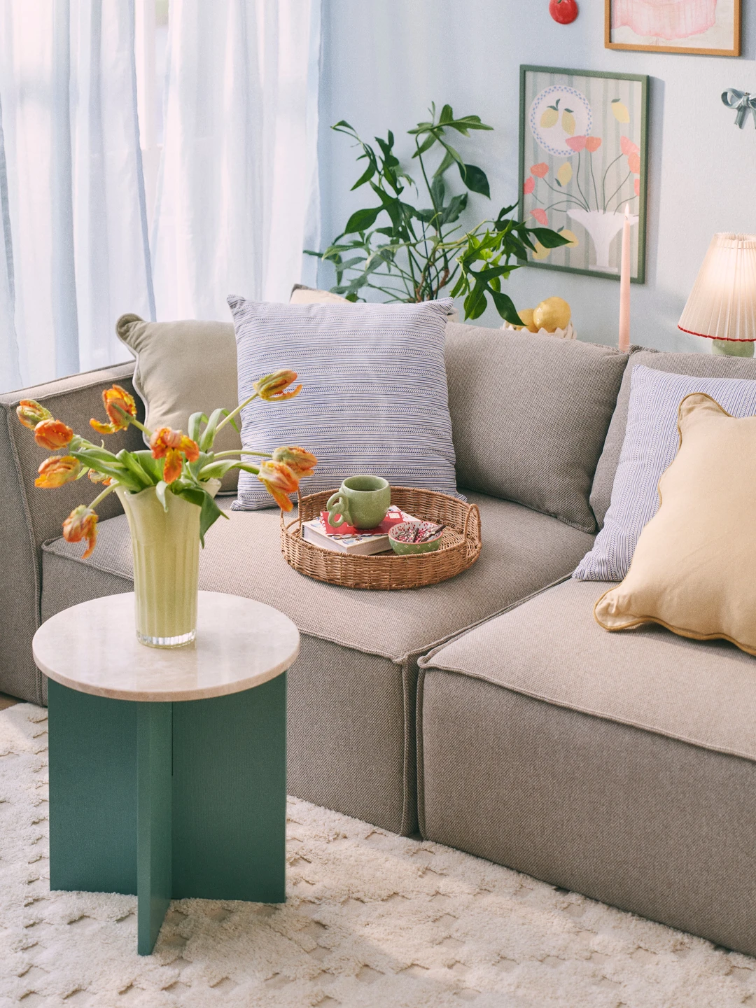 Cozy living room scene featuring a light gray sofa adorned with decorative pillows, a round green side table with a marble top, a vase filled with vibrant tulips, and a coffee tray with mugs and fruits, complemented by potted plants and soft curtains.