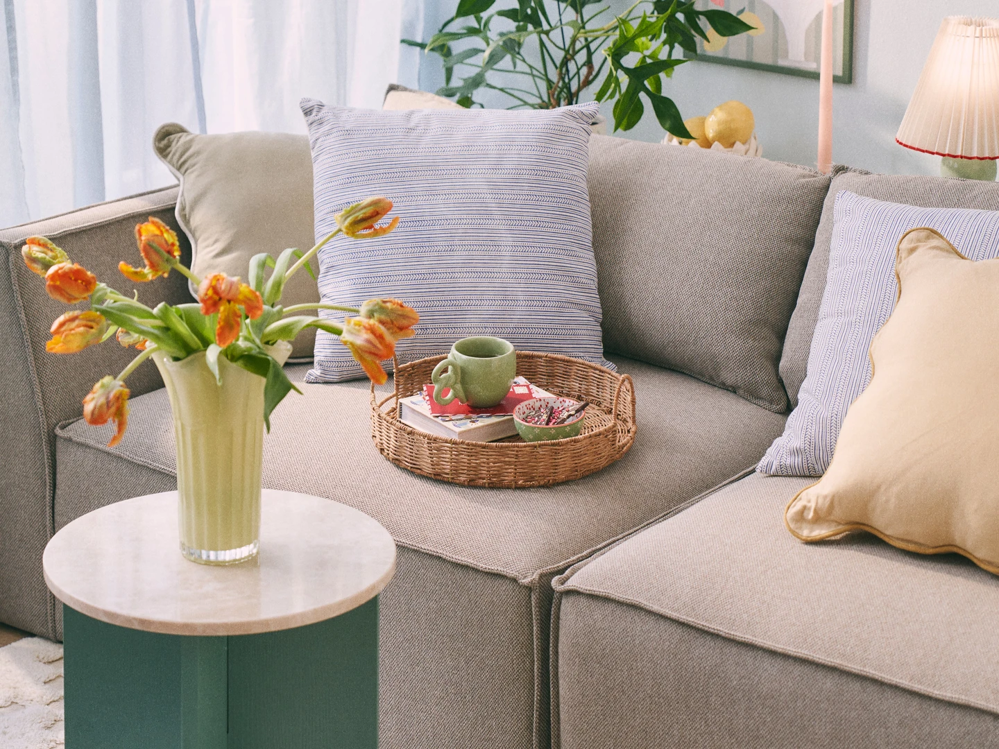 En hyggelig stue med en beige sofa med stribede puder, et rundt bord med marmorplade, en vase med tulipaner, og en kurv med frugt, omgivet af grønne planter og bløde gardiner.