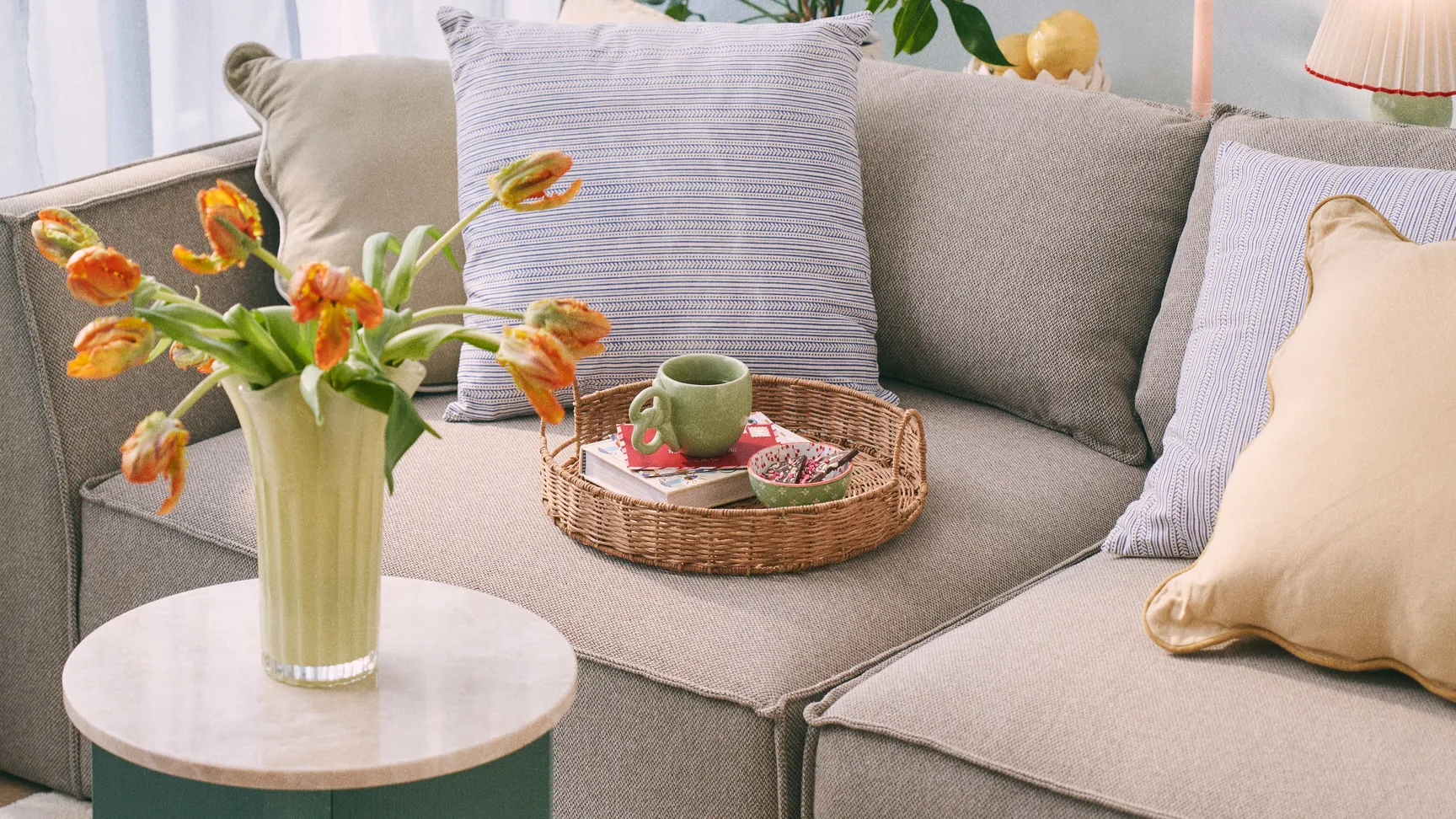 En hyggelig stue med en beige sofa med stribede puder, et rundt bord med marmorplade, en vase med tulipaner, og en kurv med frugt, omgivet af grønne planter og bløde gardiner.