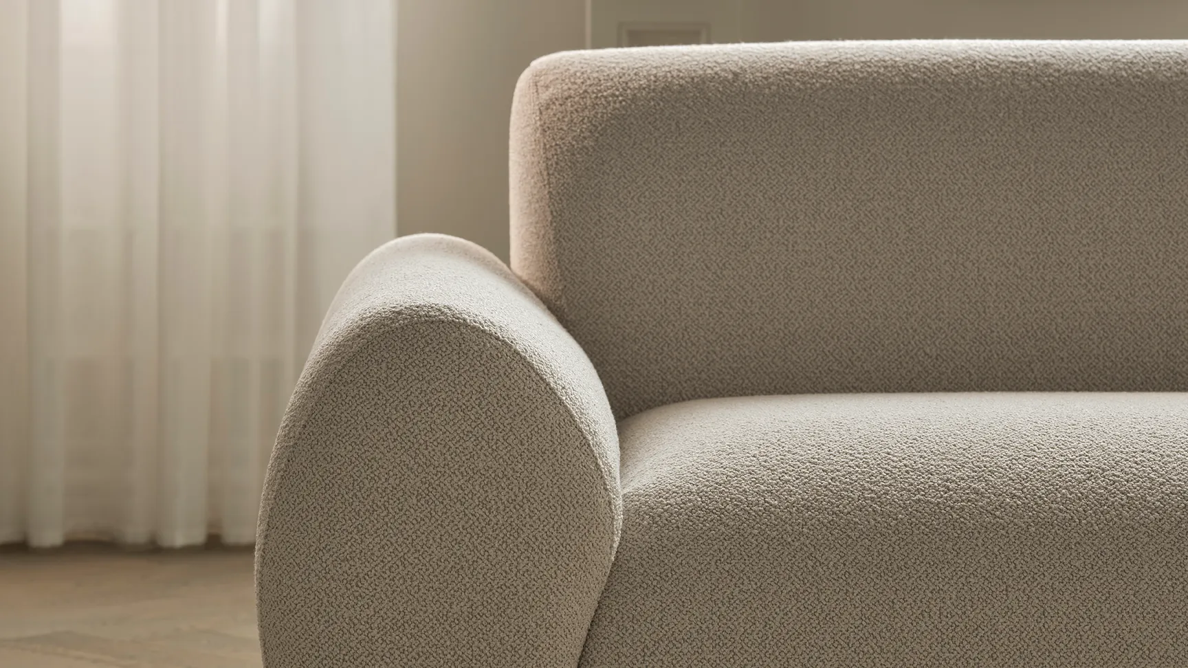 En moderne sofa med bløde kurver i en lys beige farve, placeret på et mønstrede tæppe, med lys, der filtreres gennem en hvid gardin i et stilrent rum.