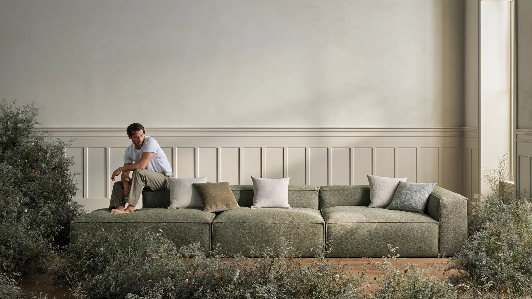 Mand sidder på en grøn sofa omgivet af bløde puder og en naturlig, vild blomsterdekor i en lys stue med enkle, lyse vægge.