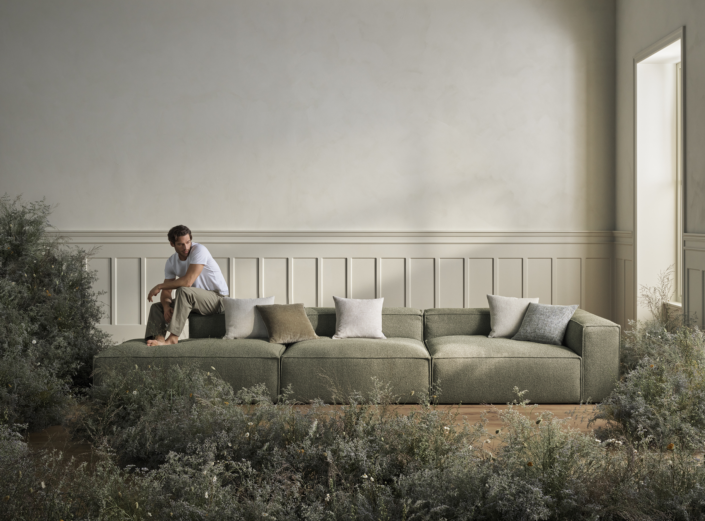 Mand sidder på en grøn sofa omgivet af bløde puder og en naturlig, vild blomsterdekor i en lys stue med enkle, lyse vægge.