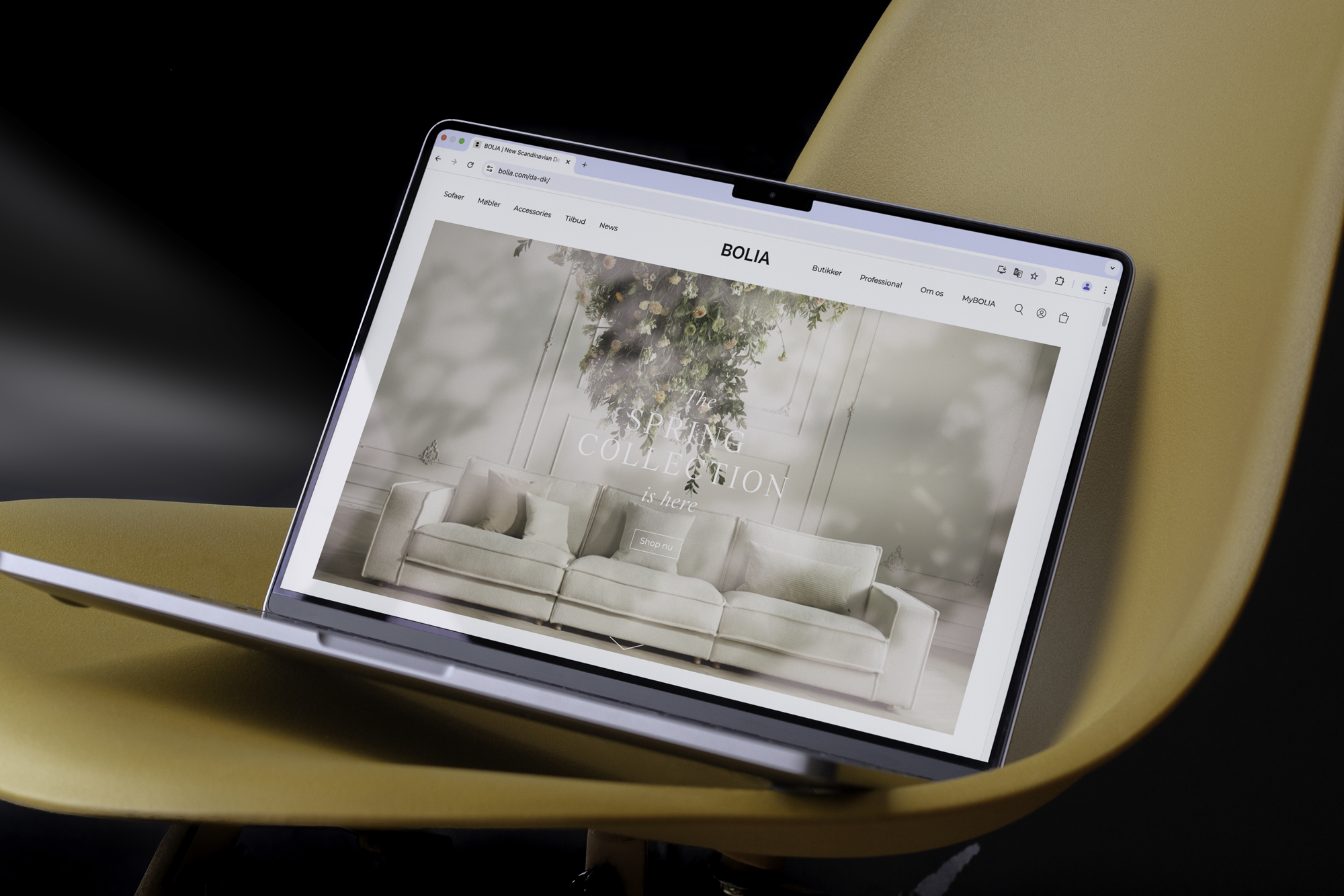 Laptop viser Bolia's hjemmeside med fokus på 'Spring Collection' og et stilrent sofa-design, placeret på en gul stol.