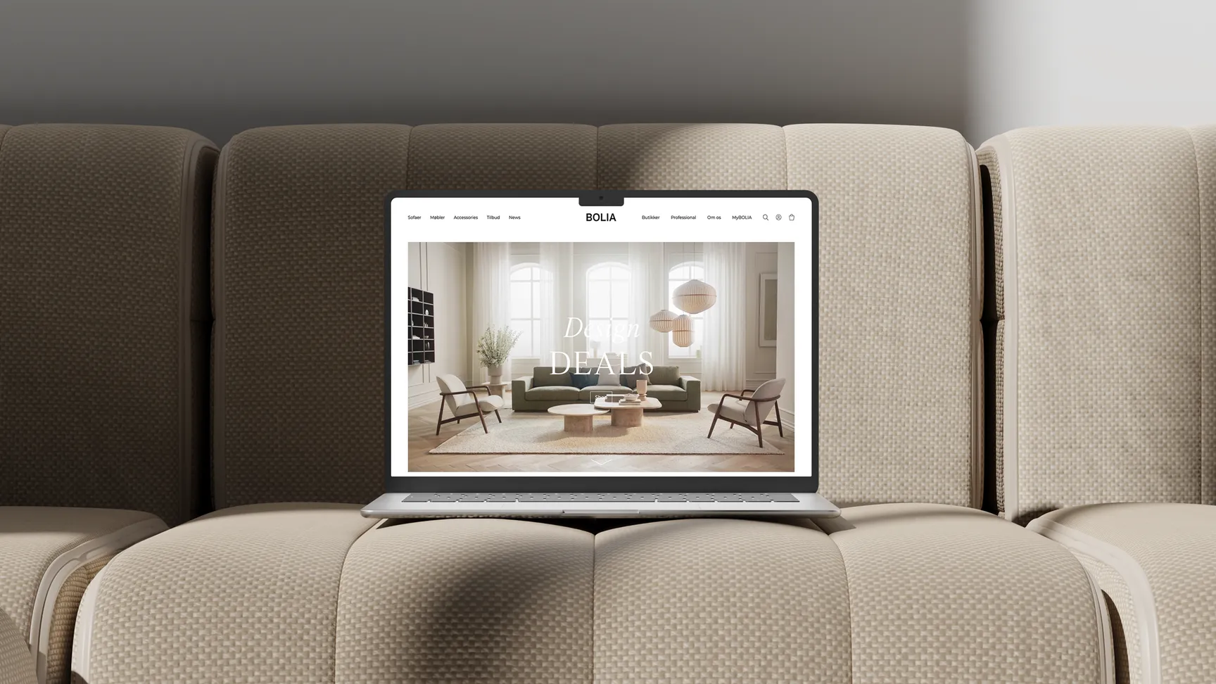 En computer står på en lys beige sofa, på computeren vises der et billede af en lys og luftig stue som er på Bolias hjemmeside