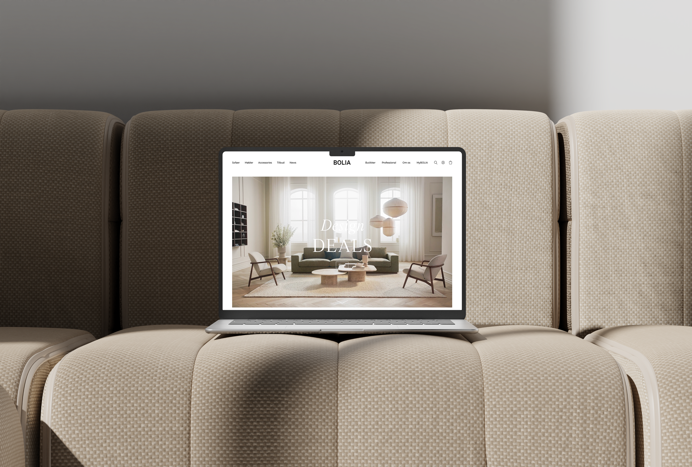 En computer  står på en lys beige sofa, på computeren vises der et billede af en lys og luftig stue som er på Bolias hjemmeside