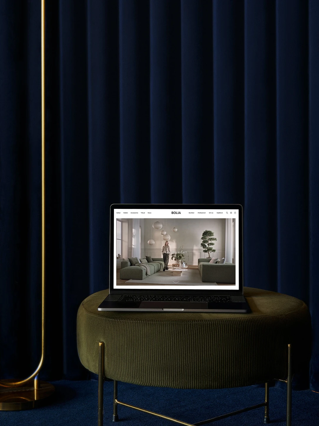 Laptop med Bolia hjemmeside, der viser et moderne stueinteriør med grønne sofaer og en plante, belyst af varme farver og omgivet af mørkeblå gardiner.