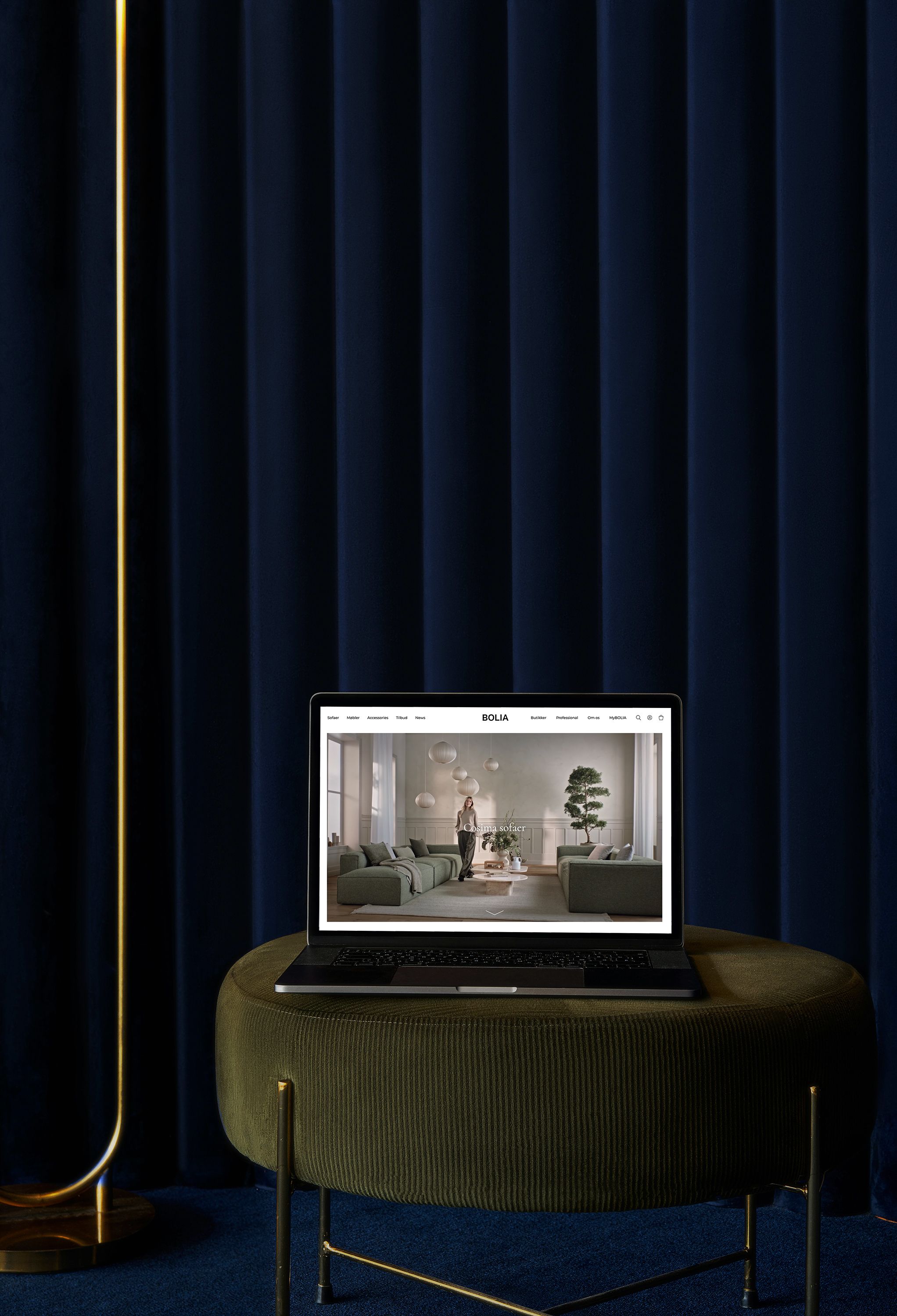 Laptop med Bolia hjemmeside, der viser et moderne stueinteriør med grønne sofaer og en plante, belyst af varme farver og omgivet af mørkeblå gardiner.