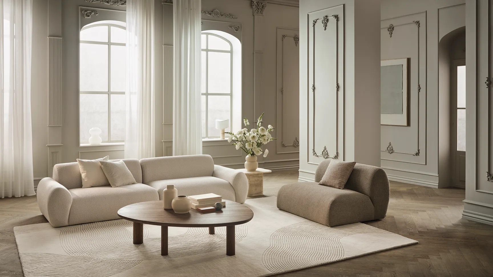 Lyst og moderne stue med stilren beige sofa, en kompakt pude, et rundt trækaffebord, og en vase med hvide blomster, omgivet af store vinduer og elegante gardiner.