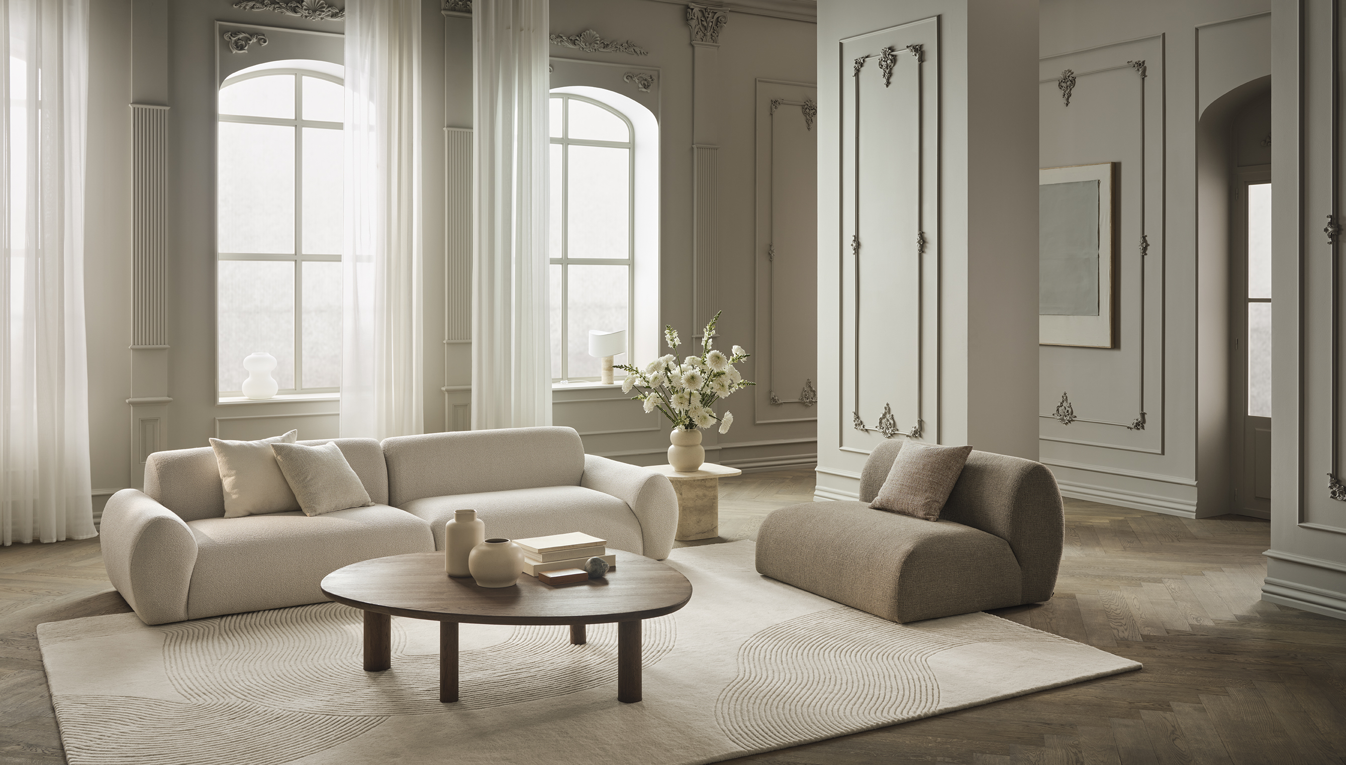 Lyst og moderne stue med stilren beige sofa, en kompakt pude, et rundt trækaffebord, og en vase med hvide blomster, omgivet af store vinduer og elegante gardiner.