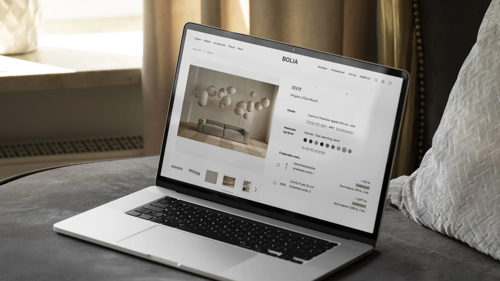 Billede af en bærbar computer, der viser Bolia's hjemmeside på en sofa omgivet af en elegant indendørs atmosfære med gardiner og puder.