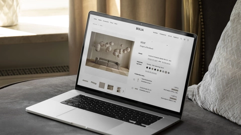 Billede af en bærbar computer, der viser Bolia's hjemmeside på en sofa omgivet af en elegant indendørs atmosfære med gardiner og puder.