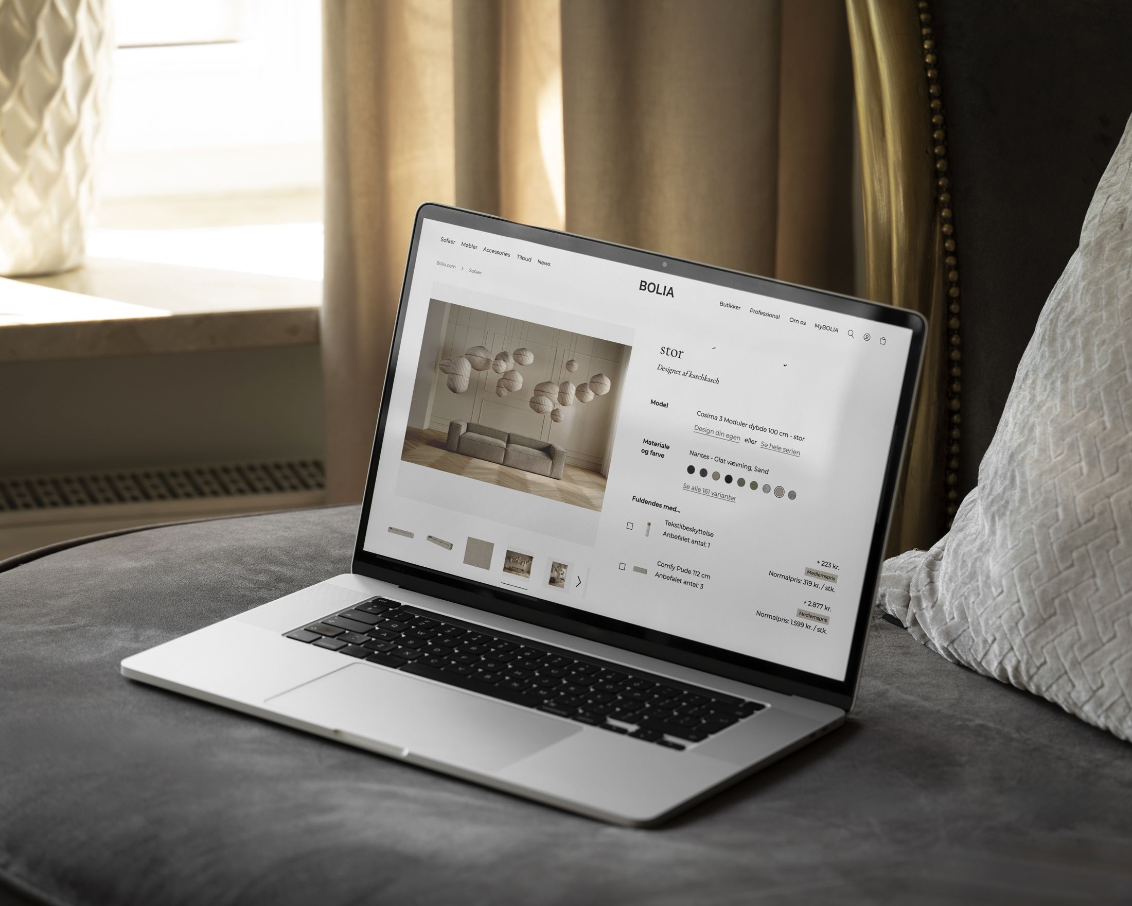 Billede af en bærbar computer, der viser Bolia's hjemmeside på en sofa omgivet af en elegant indendørs atmosfære med gardiner og puder.