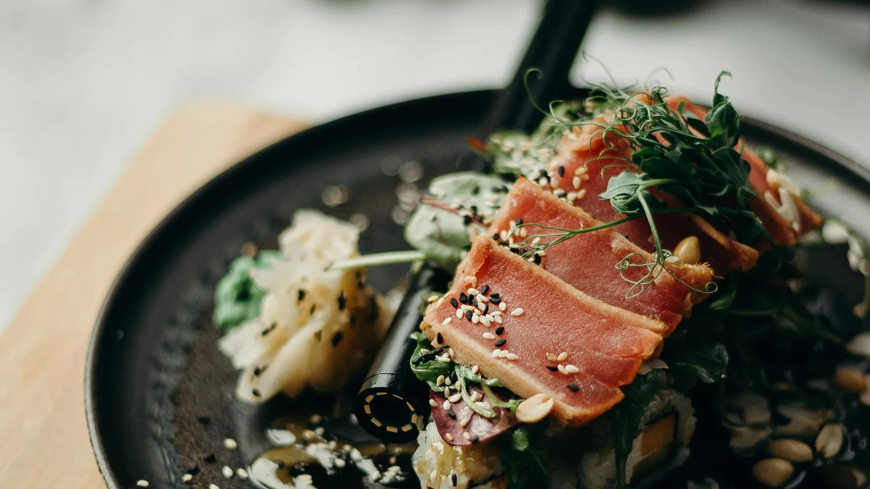 Lækker sushi på en sort tallerken med rå tun, friske urter, sesamfrø og en sojasauce, præsenteret på et træbræt.