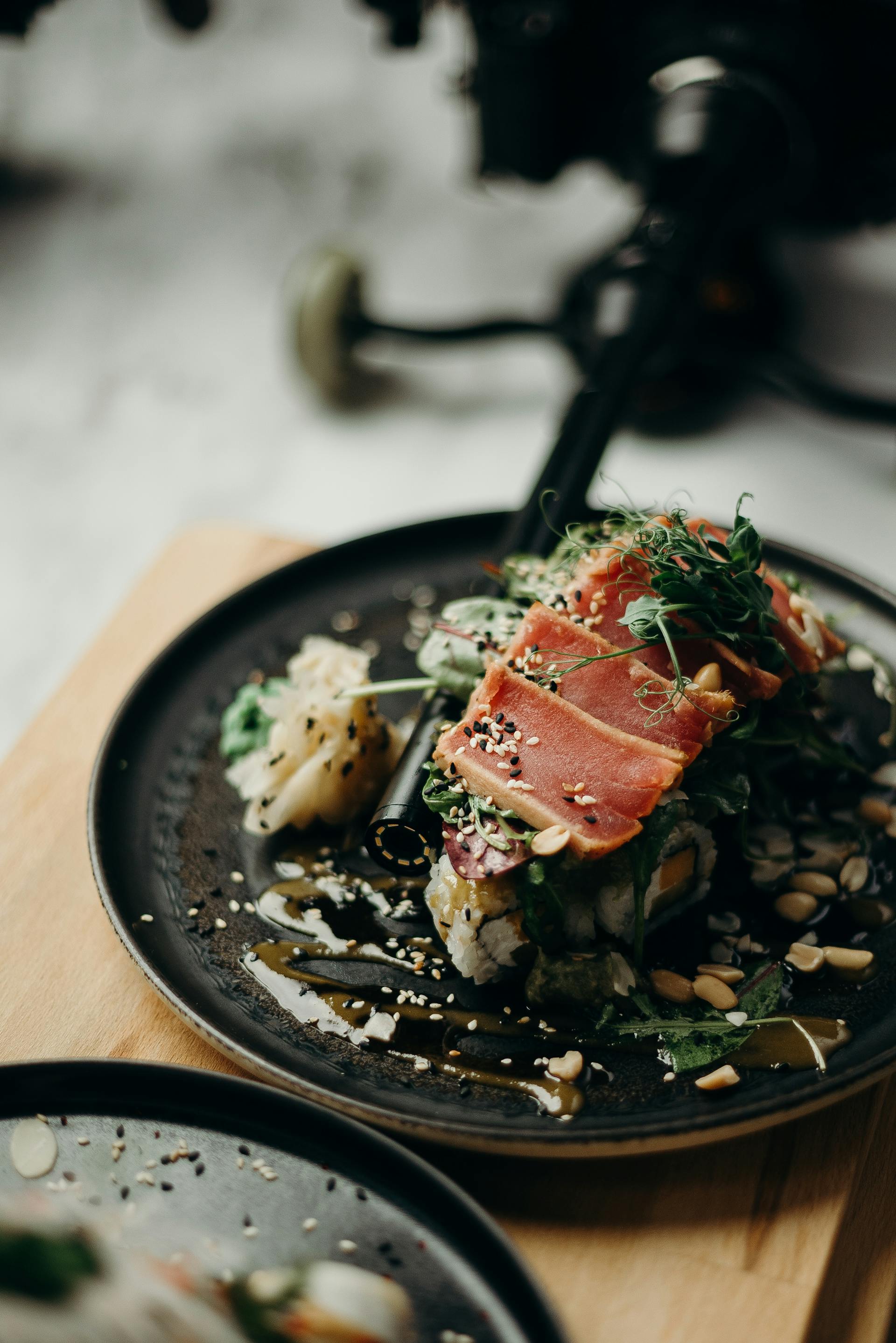 Lækker sushi på en sort tallerken med rå tun, friske urter, sesamfrø og en sojasauce, præsenteret på et træbræt.