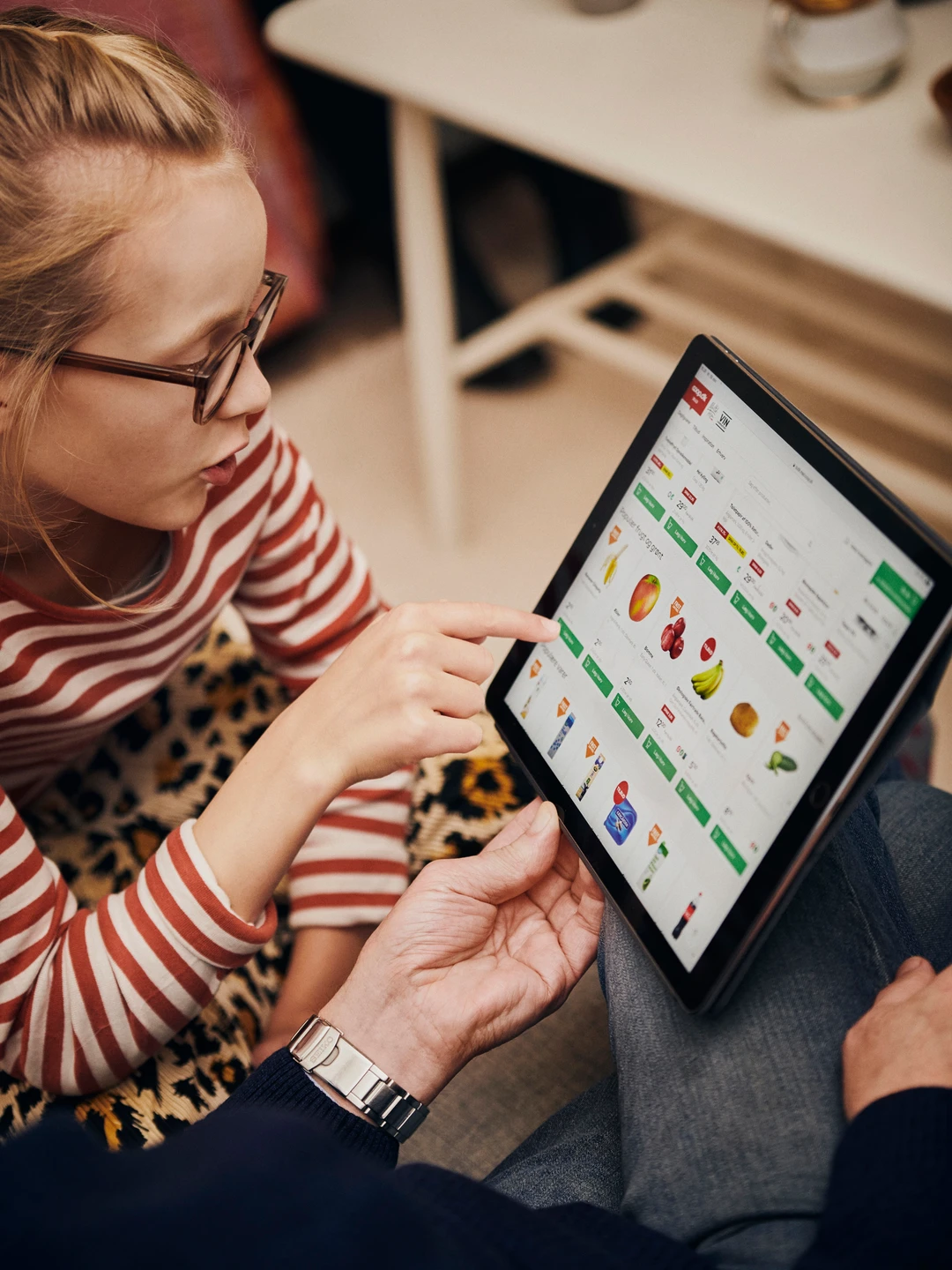 Barn peger på en tablet med madvarer, mens en voksen ser på, i et hyggeligt hjemmemiljø.