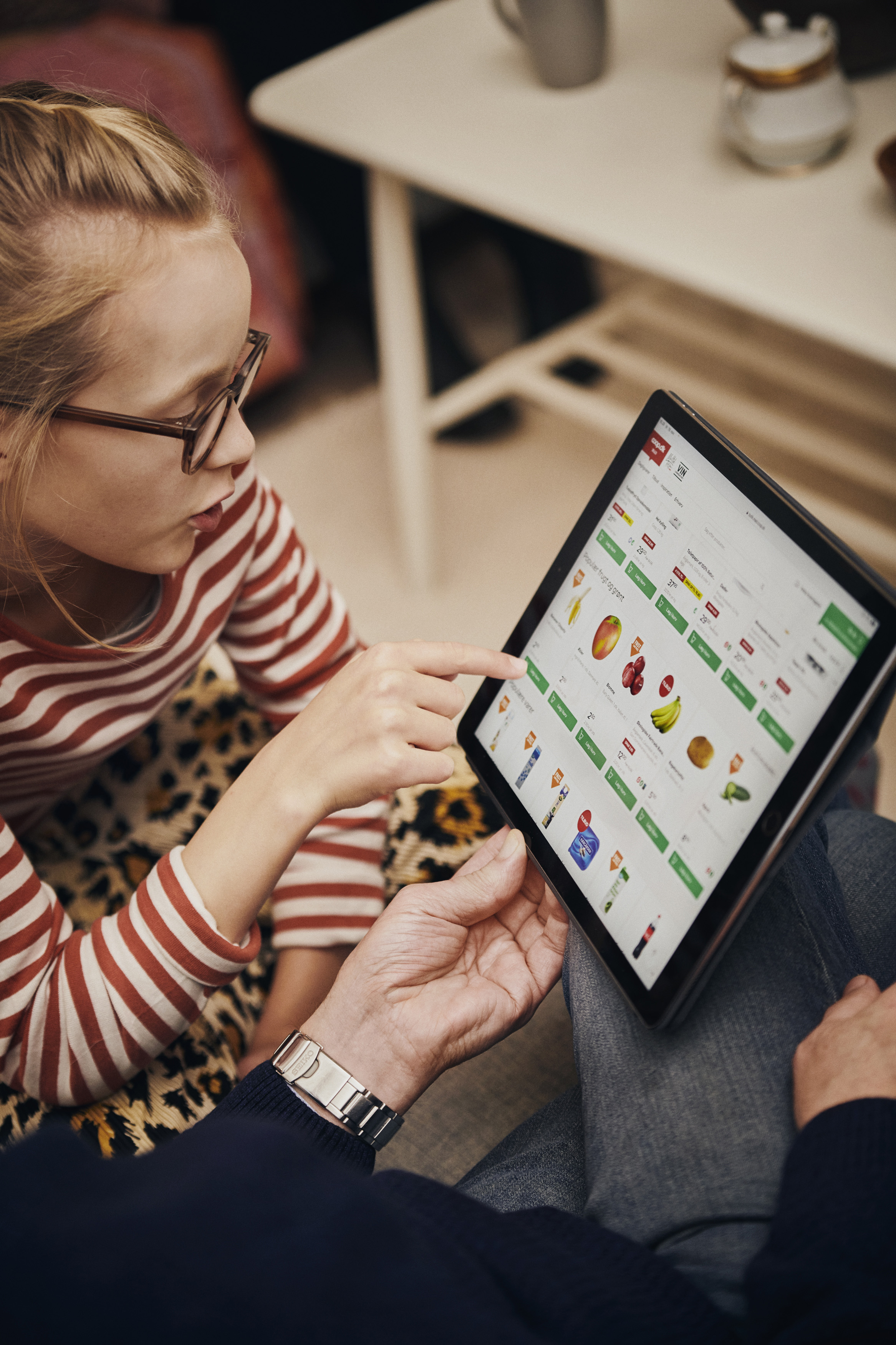 Barn peger på en tablet med madvarer, mens en voksen ser på, i et hyggeligt hjemmemiljø.