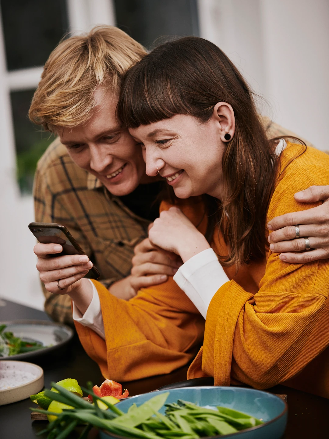 Et par, der smiler og kigger på en telefon, mens de sidder ved et bord dækket med friske grøntsager og salater.