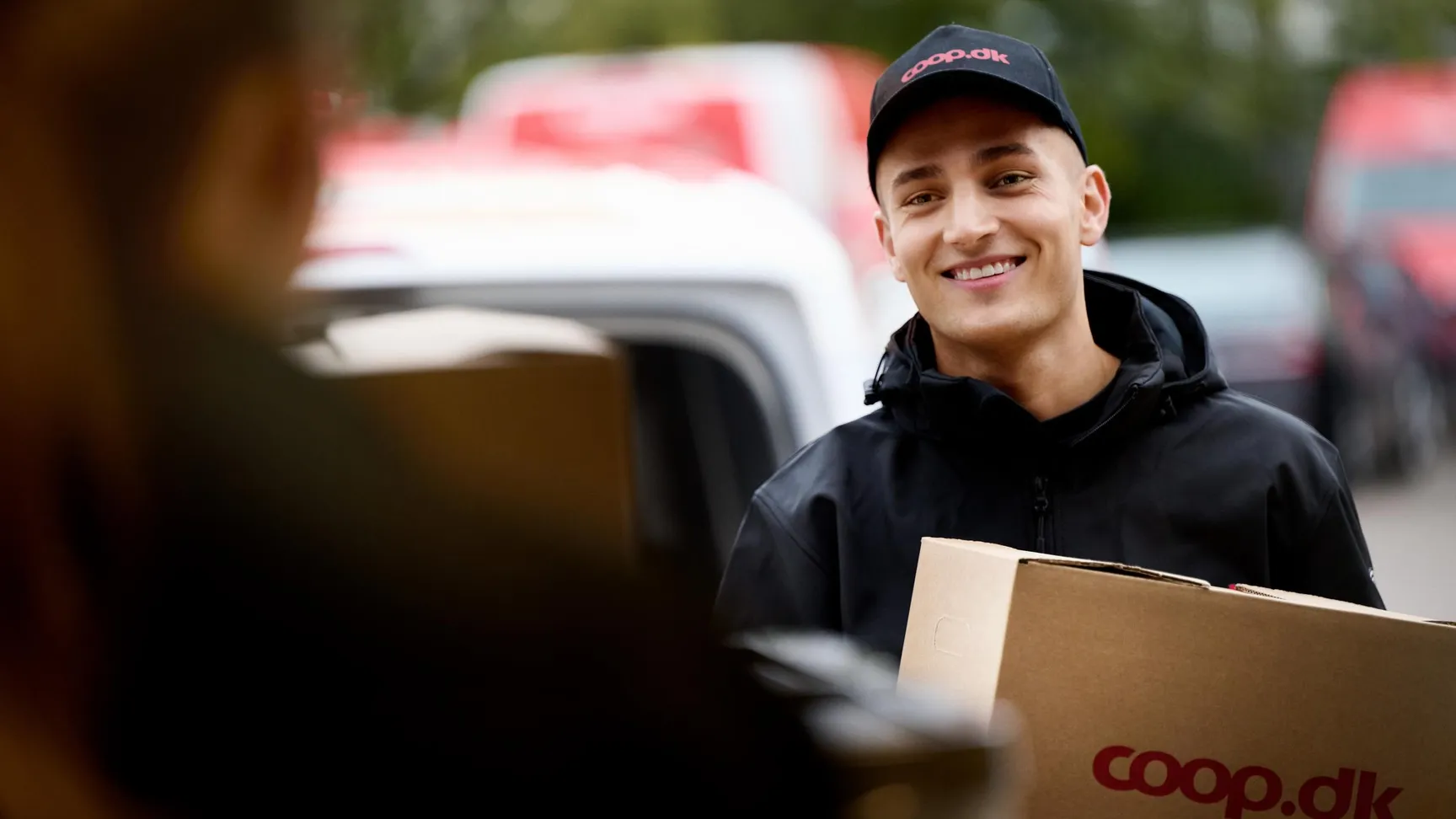 Ung mand med smugende smil bærer en kasse fra coop.dk, mens han interagerer med en kunde, med bil og leveringsmiljø i baggrunden.