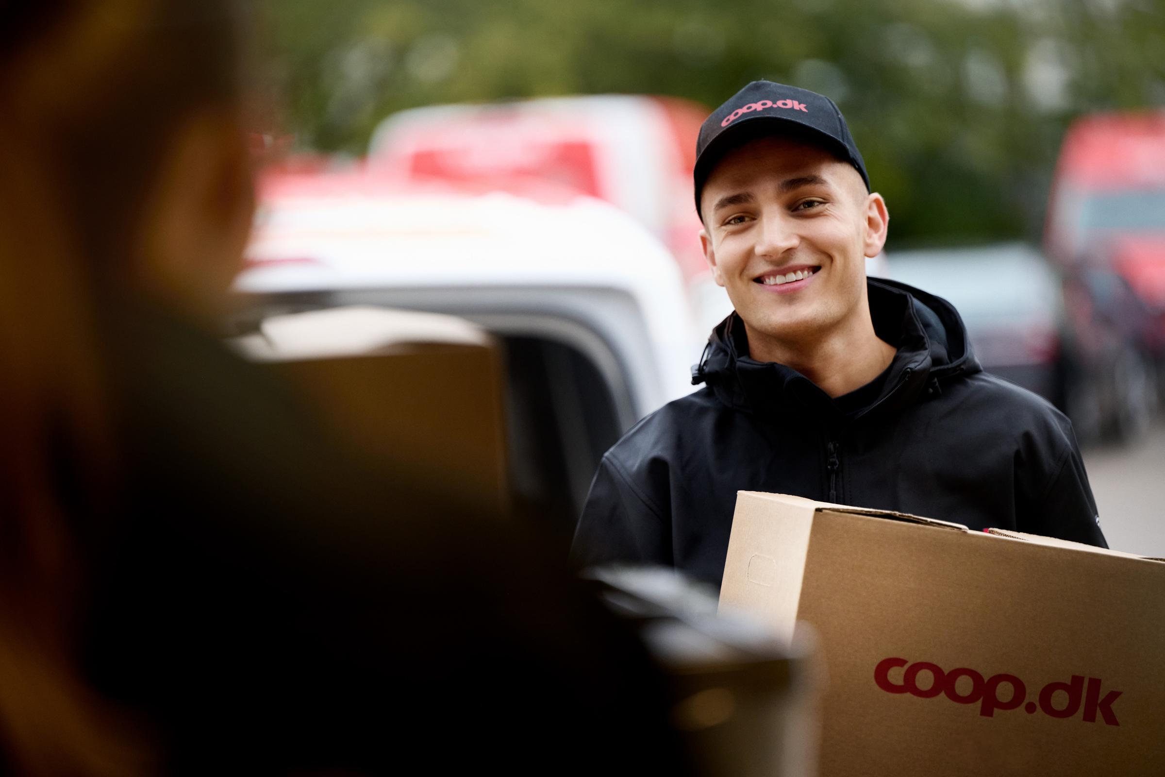 Ung mand med smugende smil bærer en kasse fra coop.dk, mens han interagerer med en kunde, med bil og leveringsmiljø i baggrunden.