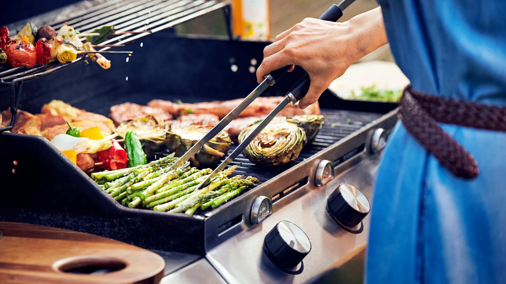 En person griller asparges og grøntsager på en gasgrill, med fokus på maden og grillens kontrolknapper, mens damp stiger op i baggrunden.
