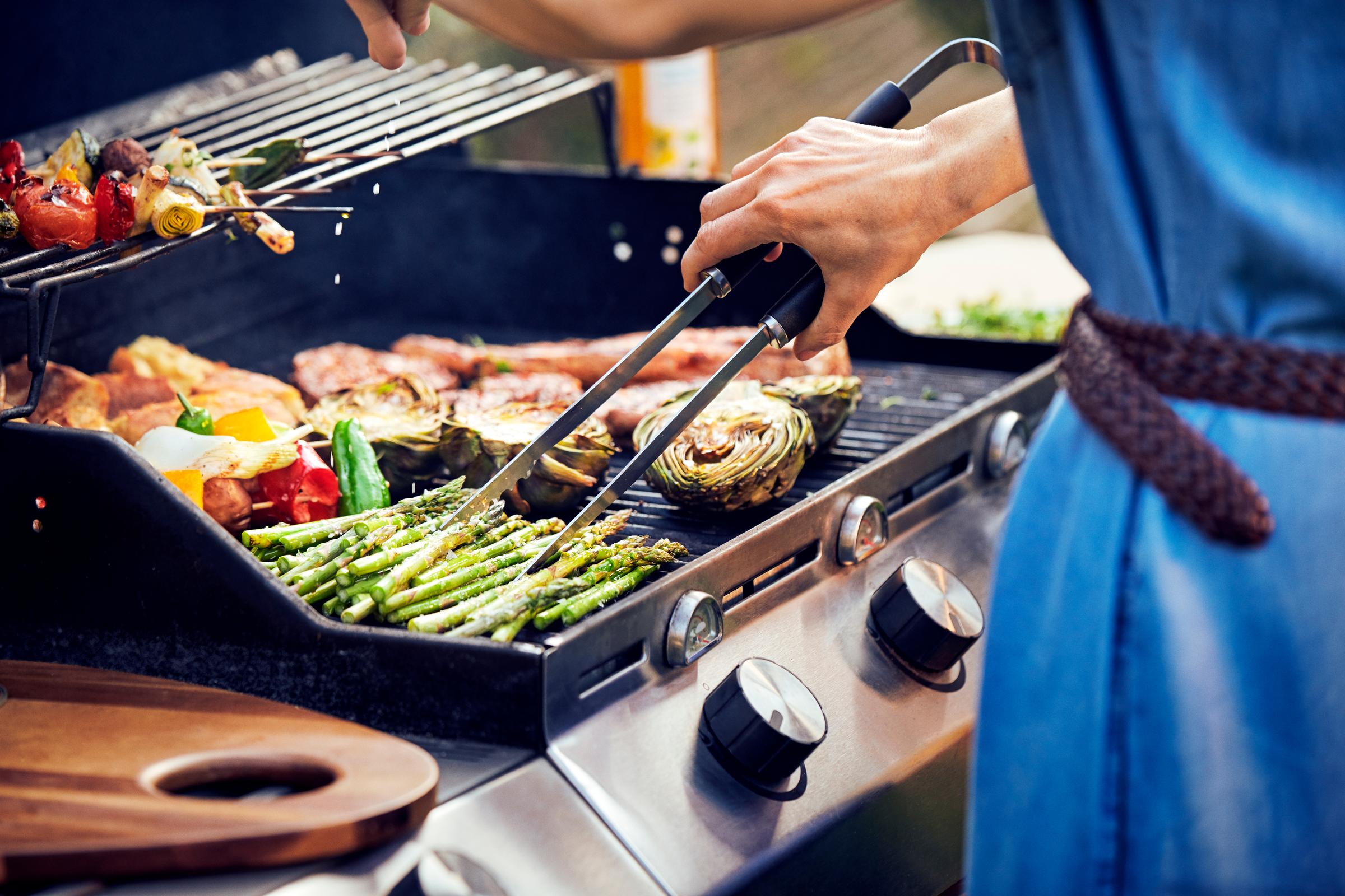 En person griller asparges og grøntsager på en gasgrill, med fokus på maden og grillens kontrolknapper, mens damp stiger op i baggrunden.