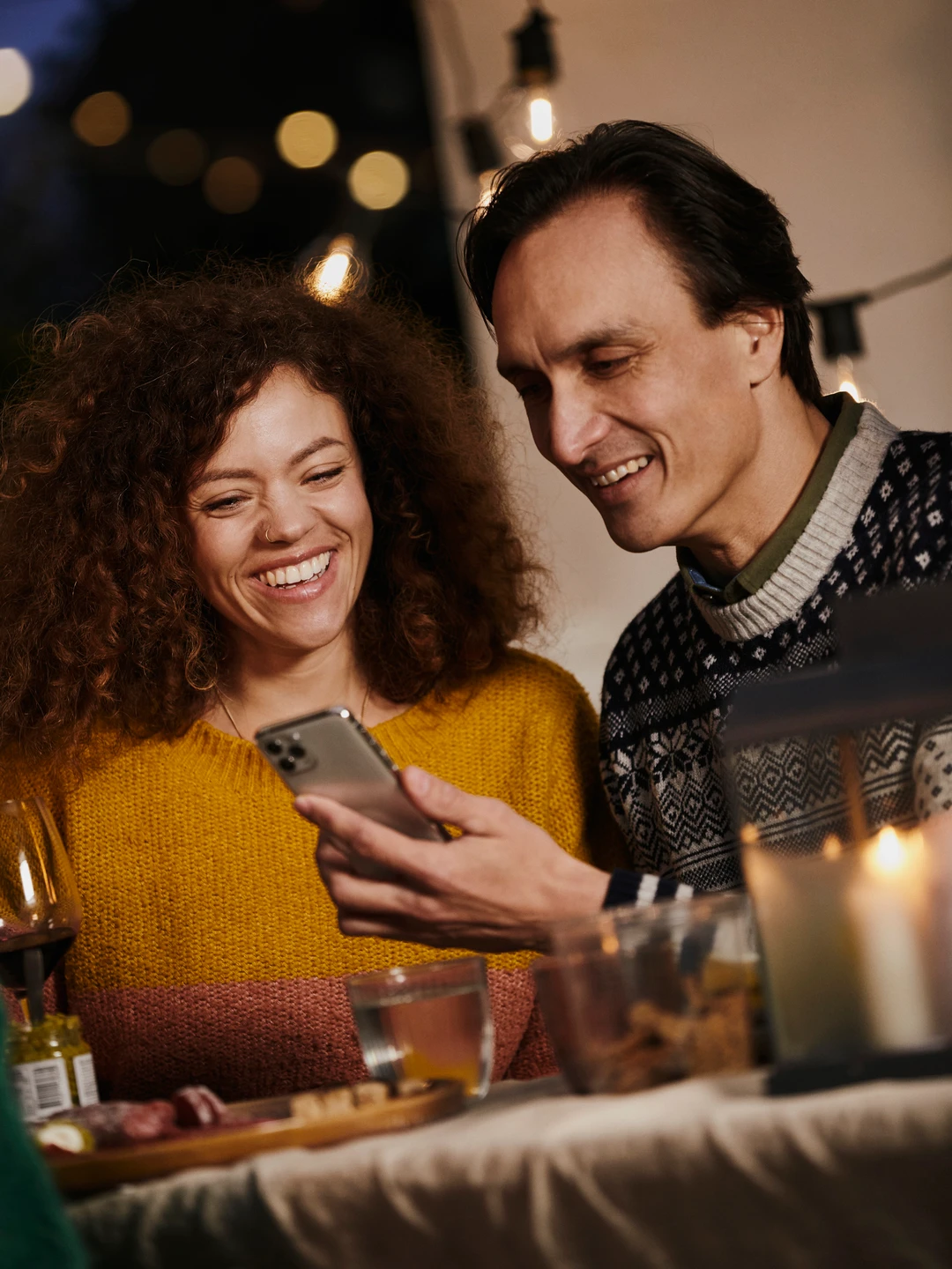 To personer, en mand og en kvinde, sidder ved et bord med mad og drikke, mens de smiler og ser på en smartphone, omgivet af blødt lys fra lyskæder.