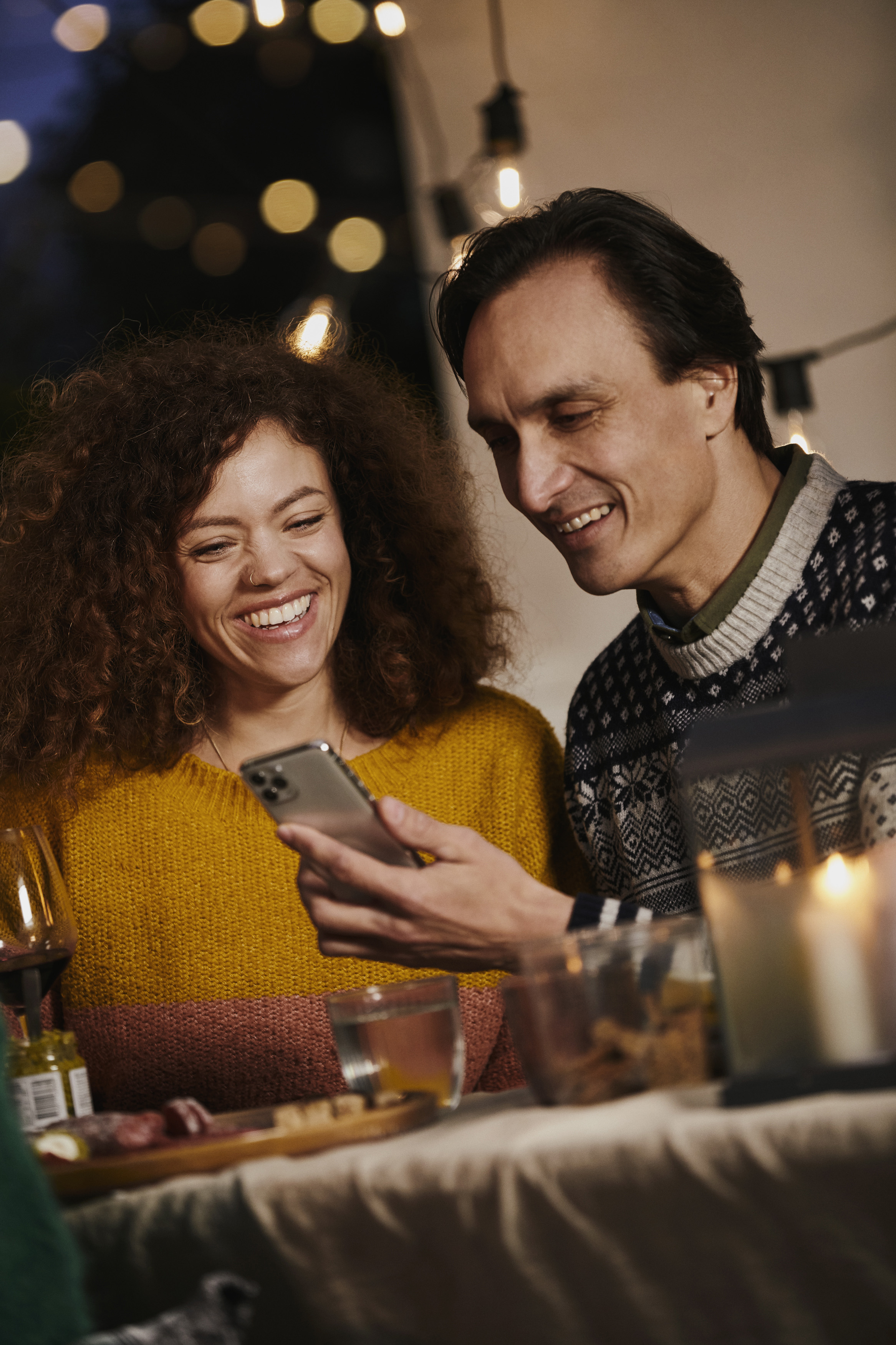 To personer, en mand og en kvinde, sidder ved et bord med mad og drikke, mens de smiler og ser på en smartphone, omgivet af blødt lys fra lyskæder.