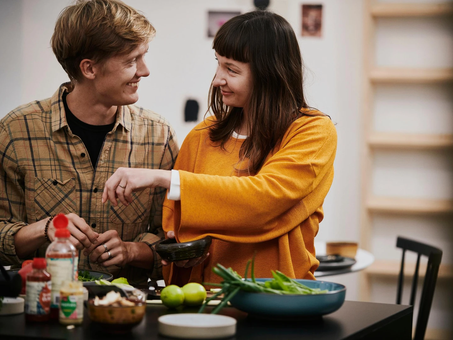 To personer smiler og interagerer over et køkkenbord fyldt med grøntsager og ingredienser, mens en af dem holder en skål.
