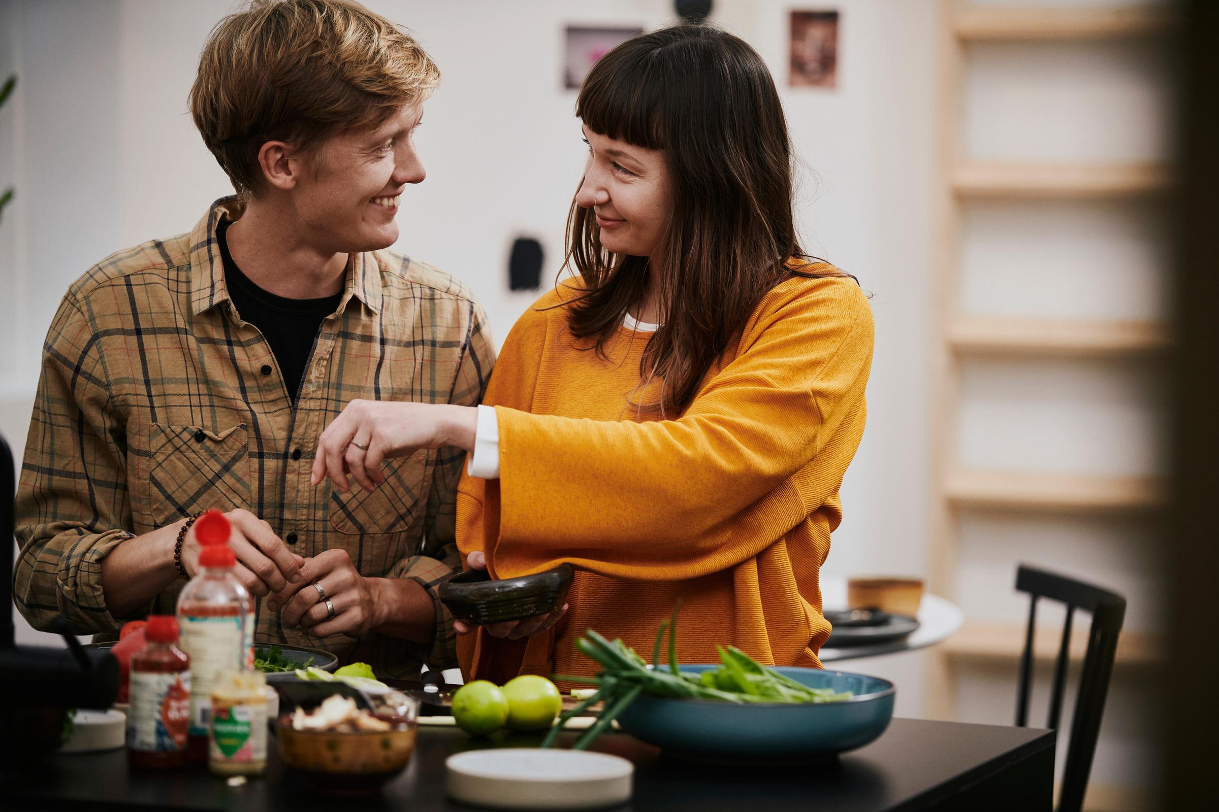 To personer smiler og interagerer over et køkkenbord fyldt med grøntsager og ingredienser, mens en af dem holder en skål.