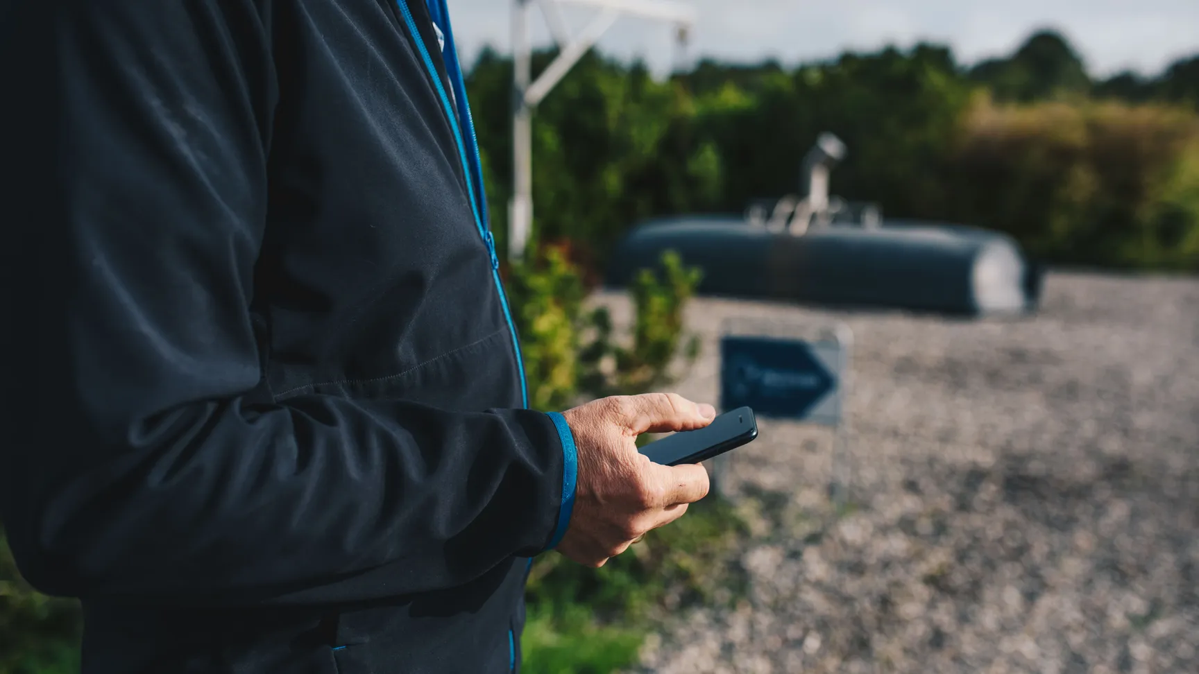 A person wearing a black jacket with blue accents holds a smartphone while standing outdoors in a gravel area with greenery and equipment in the background.