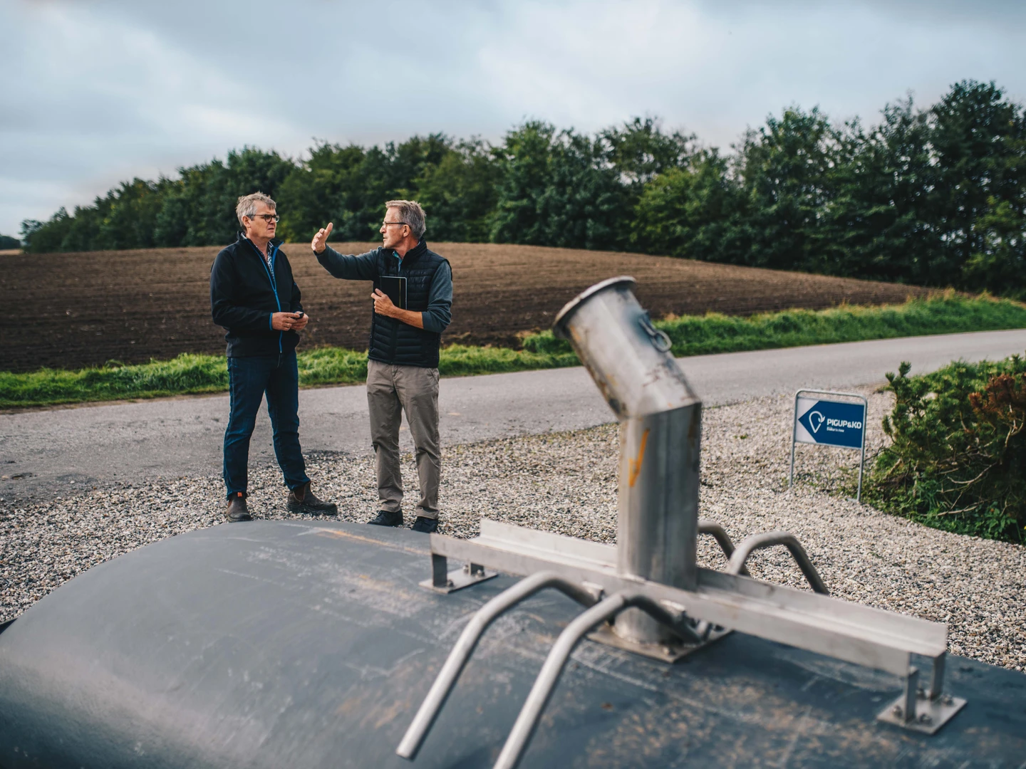 Two men discussing near a large black tank on a gravel road, with a backdrop of green trees and an agricultural field. A blue sign with a pig symbol is visible in the background.