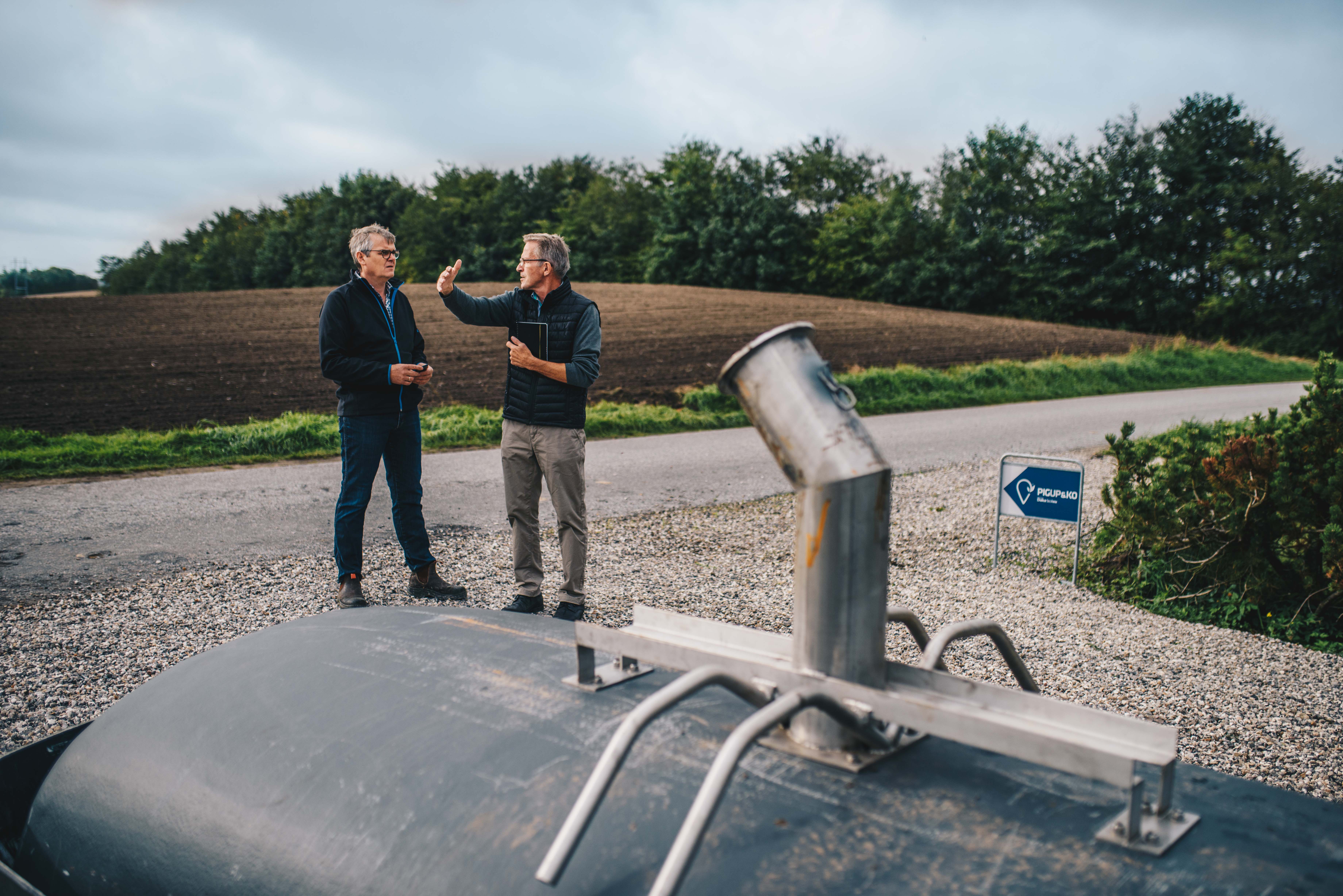 To mænd står ved en vej og diskuterer, mens de kigger på et stort, gråt metalobjekt i forgrunden; i baggrunden ses marker og træer under en overskyet himmel.