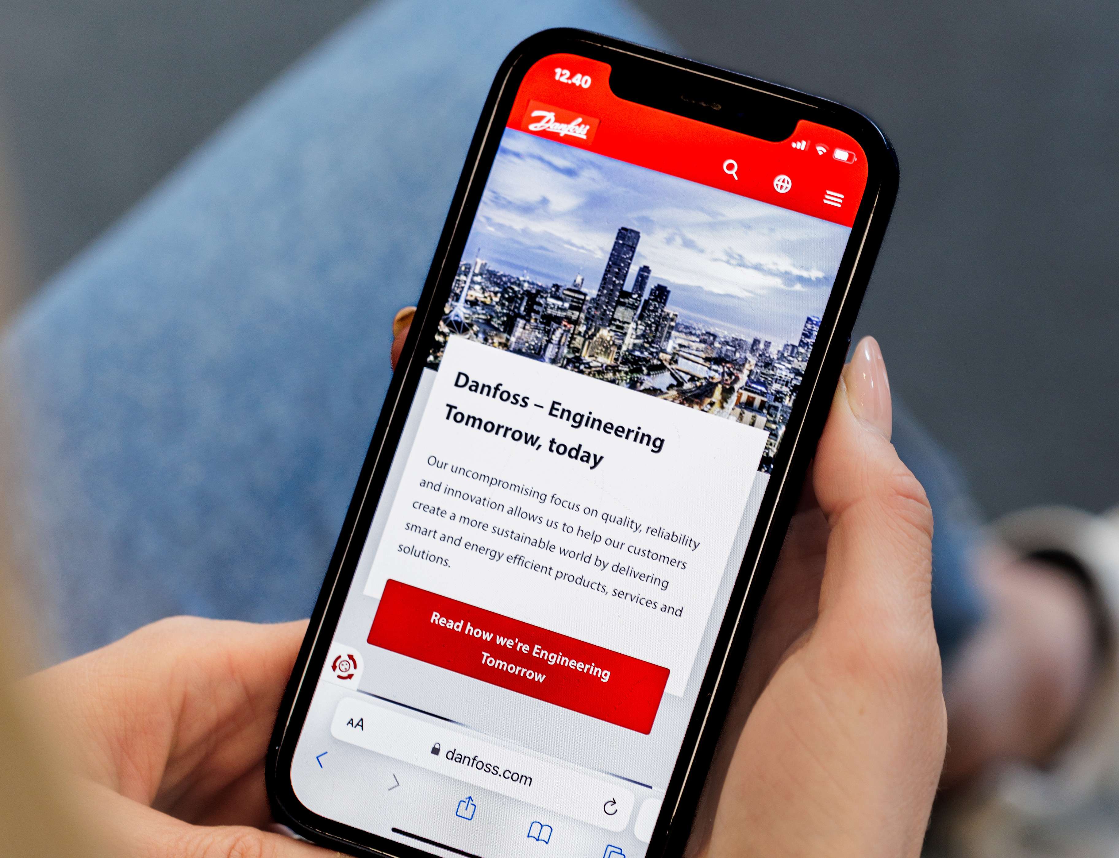 En person holder en smartphone, der viser Danfoss' hjemmeside med teksten "Danfoss - Engineering Tomorrow, today" og en bysilhuet i baggrunden.