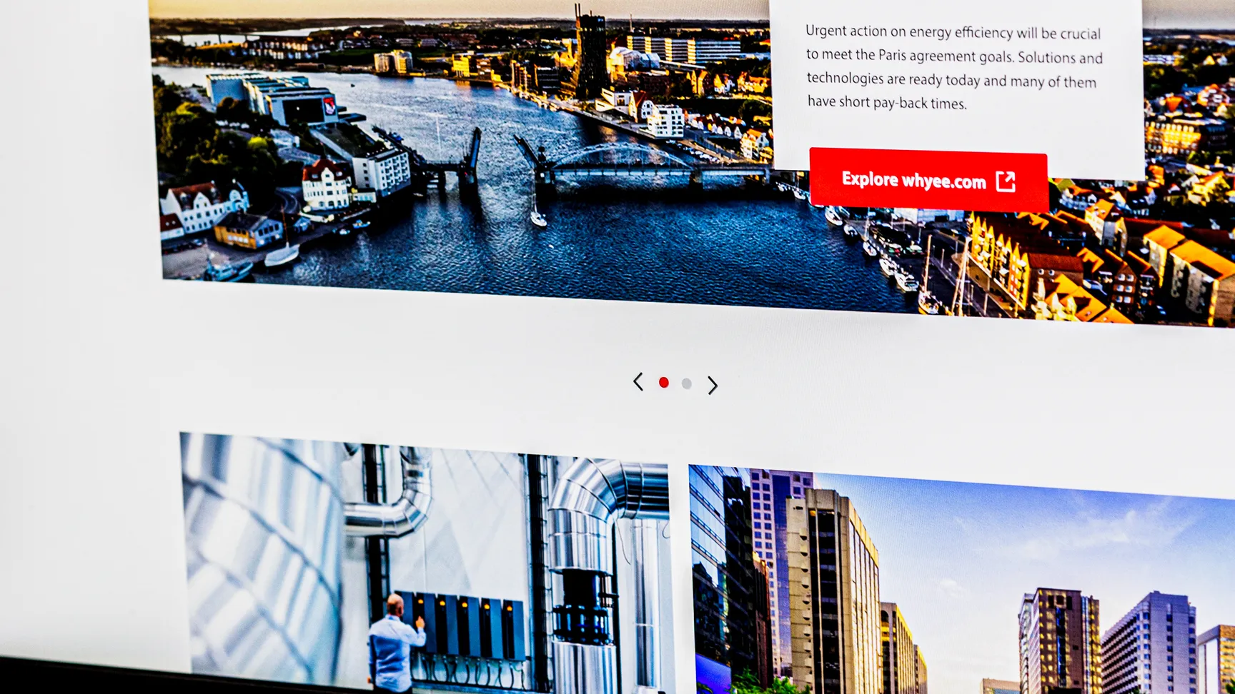 A close-up of a computer monitor displaying the Danfoss website with information about energy efficiency along with a scenic image of a river and city in the background, featuring a navigation menu at the top and a vibrant workspace setting.