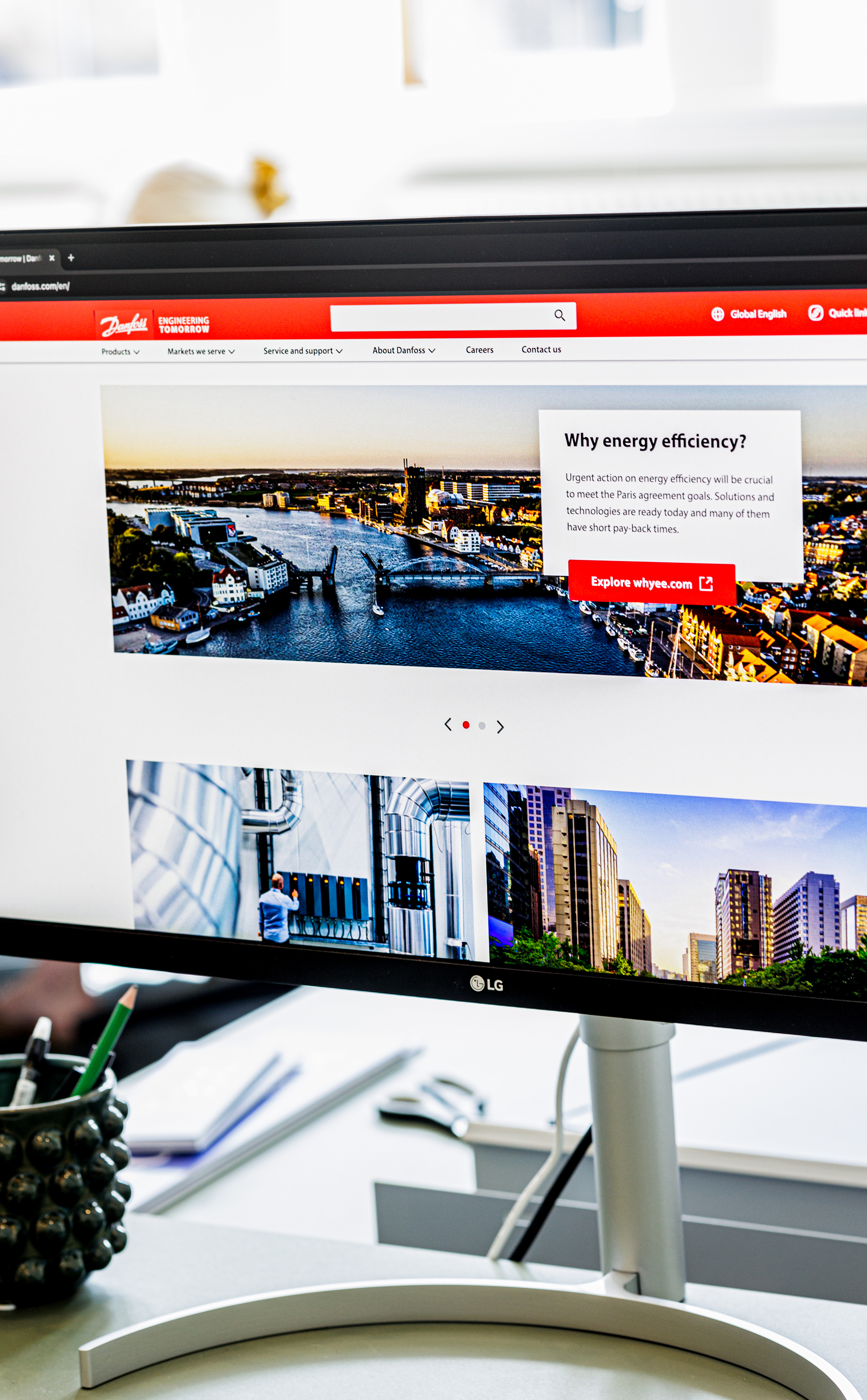 Skærmbillede af Danfoss hjemmeside, der viser information om energibesparelse med bylandskab og skridtlister på skærmen, med en penneholder og papir i forgrunden.