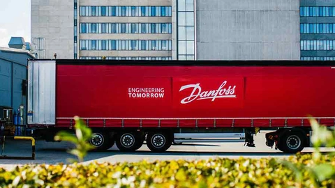 Rød lastbil med Danfoss-logo parkeret ved bygning E20, omgivet af grønne buske og moderne bygninger i baggrunden.