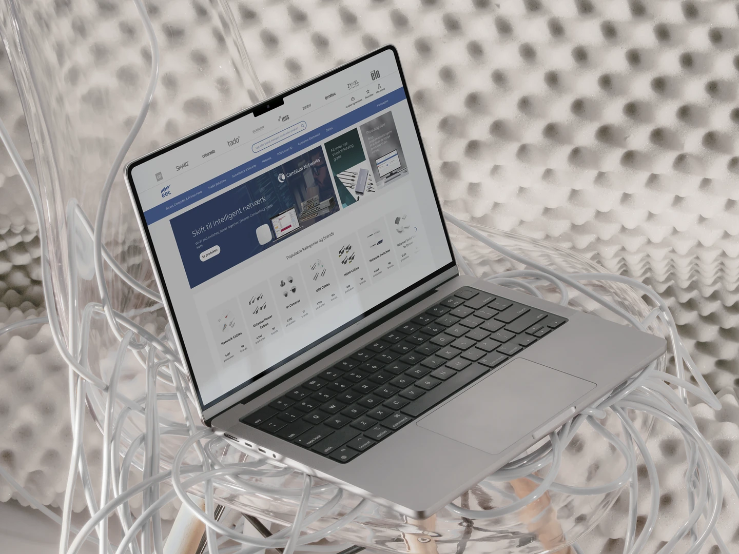 A MacBook laptop displaying a website on intelligent networking, surrounded by tangled cables, resting in a transparent chair with a textured background.